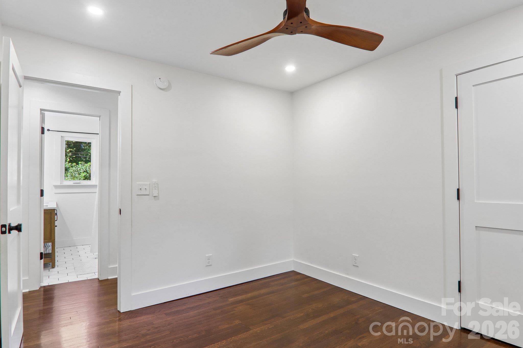 Empty bedroom with dark hardwood floors, a wood-blade ceiling fan, and an open doorway to a tiled bathroom.