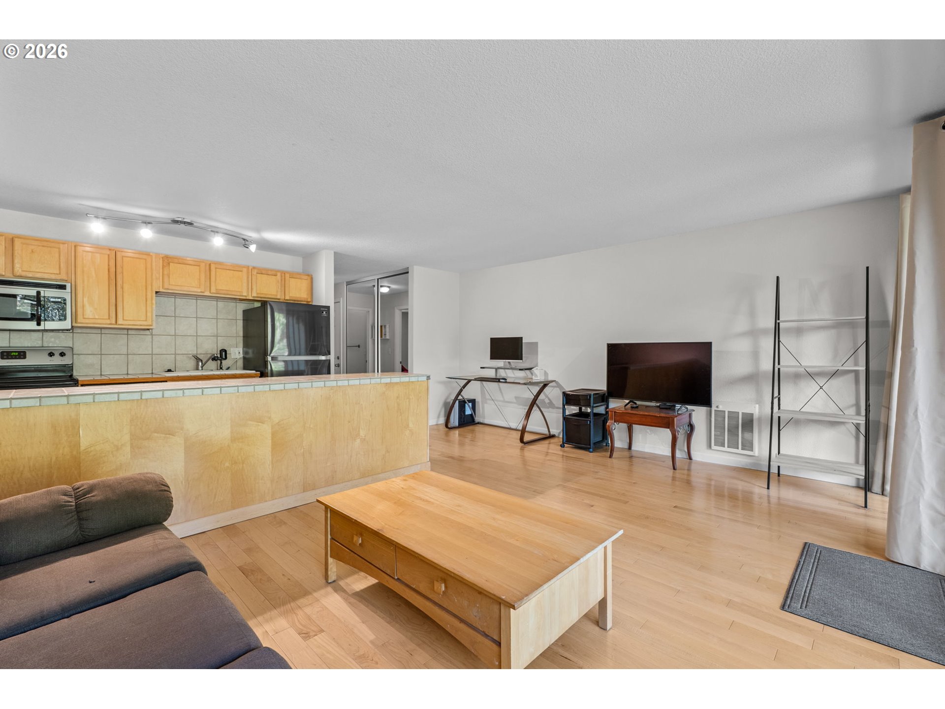Open-plan living room and kitchen with a tiled peninsula, light wood floors, and gray sofa.