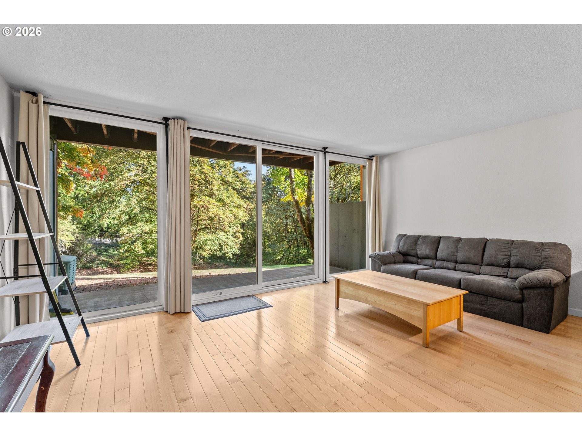 Living room with a gray sectional and wood floors, opening through sliding glass doors to a leafy patio.