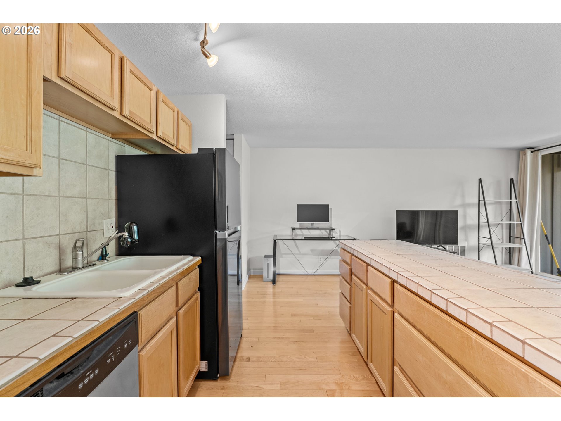Kitchen with light wood cabinets, tiled counters and a long island opening into a living area.