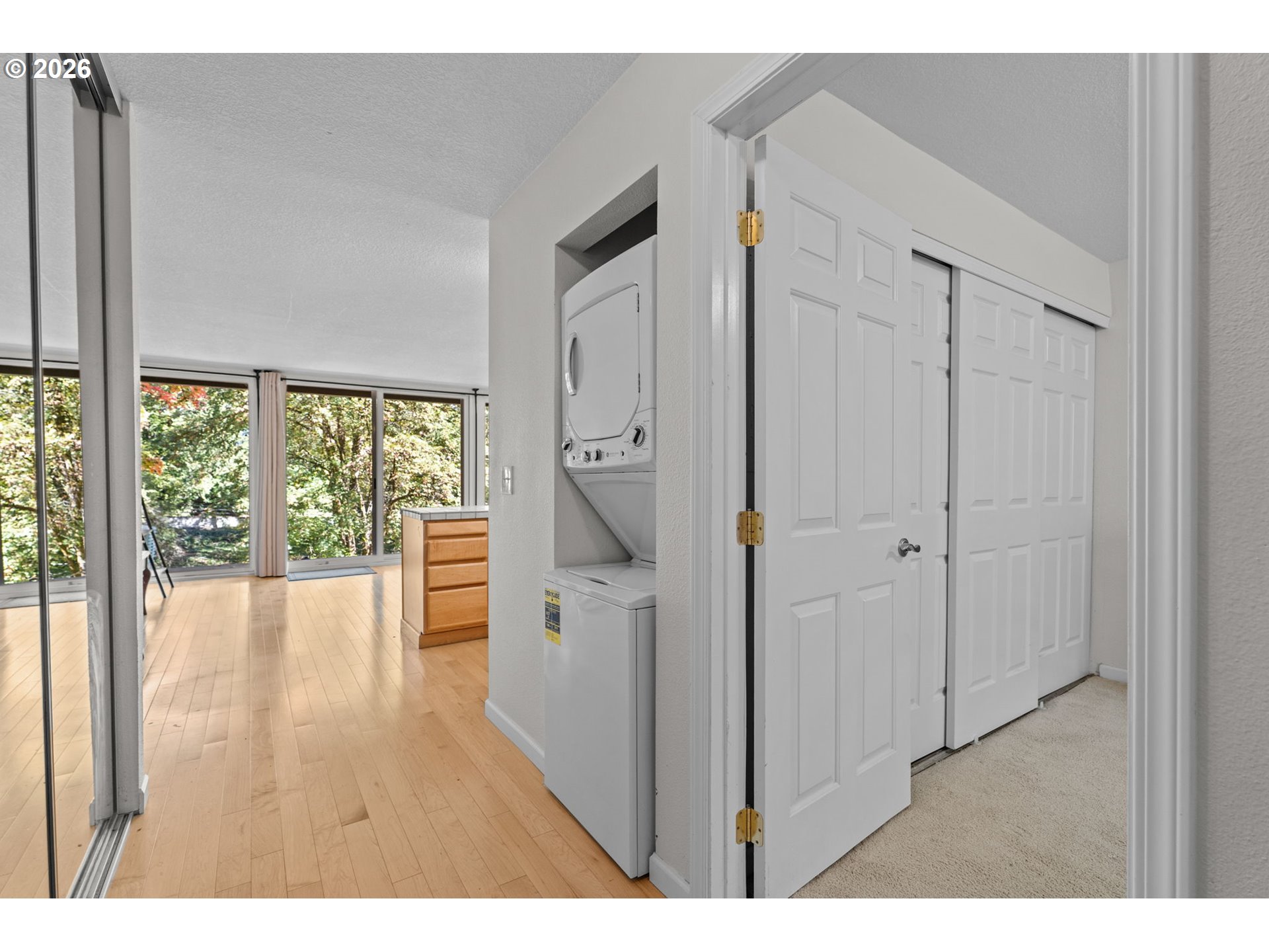 Laundry area with stacked washer and dryer, open closet doors, and large windows overlooking trees.