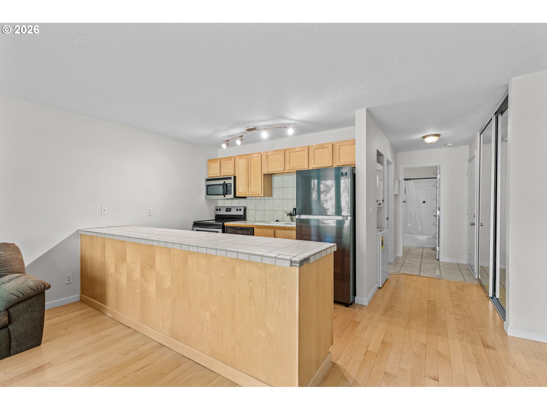 Condo kitchen with light wood cabinets, stainless steel appliances, and a tiled breakfast bar beside a hallway.