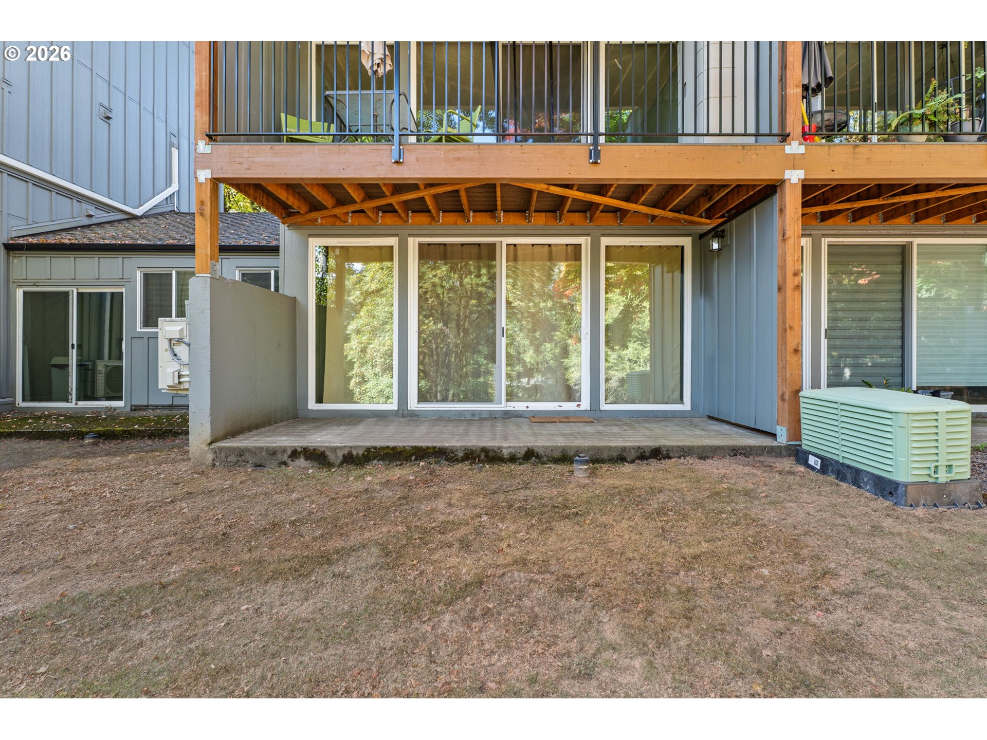 Home’s covered rear patio beneath a raised wooden deck, with sliding glass doors and a lawn.