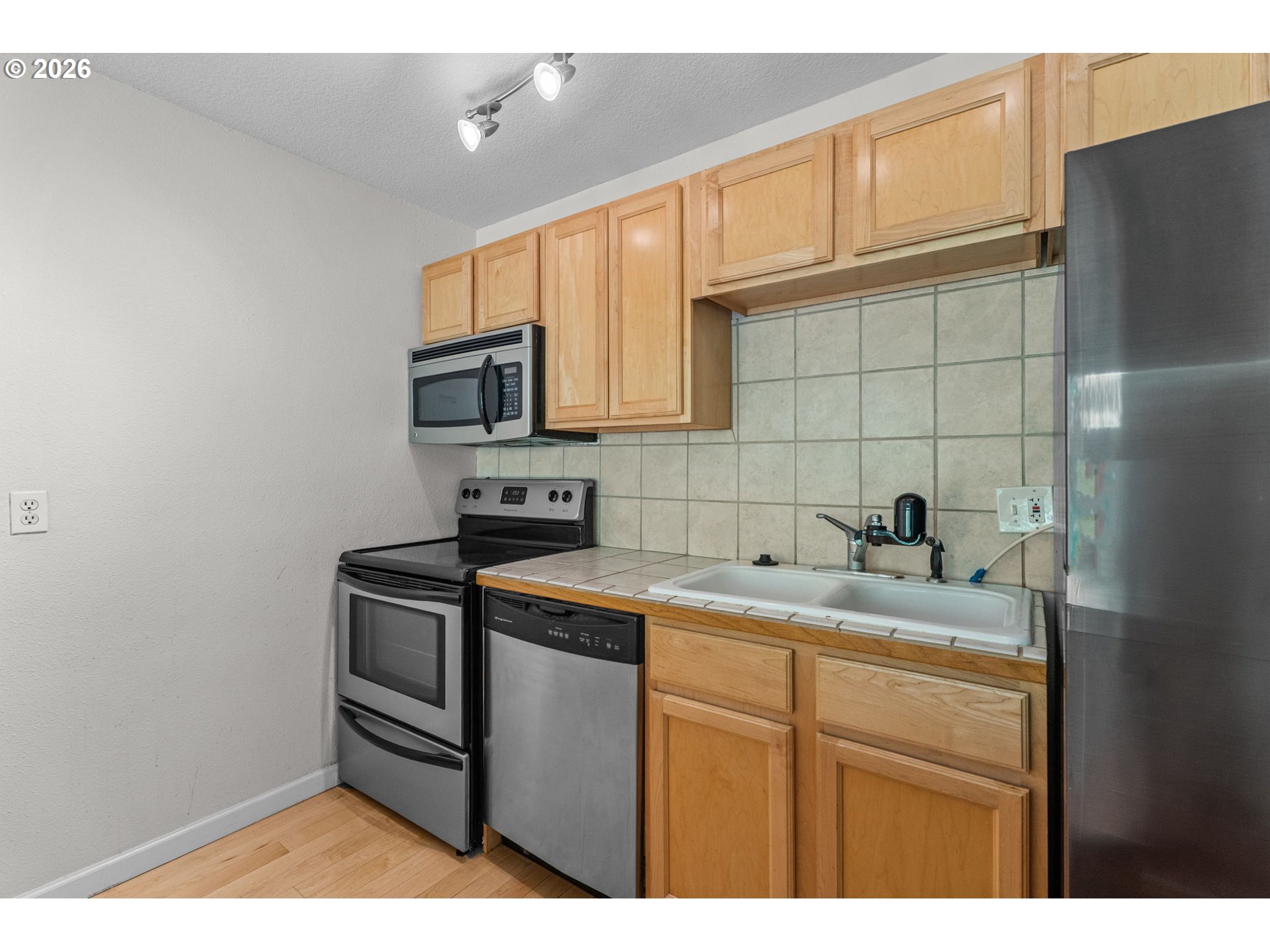 Kitchen with light wood cabinets, tiled backsplash, stainless steel appliances, and a white sink.