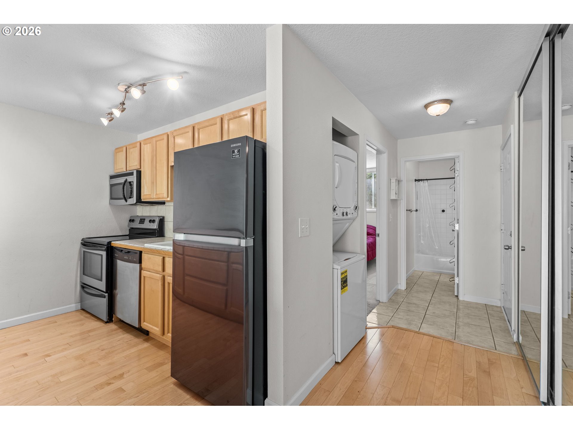 Kitchen with light wood cabinets and dark refrigerator, opening to a laundry nook and tiled bathroom.