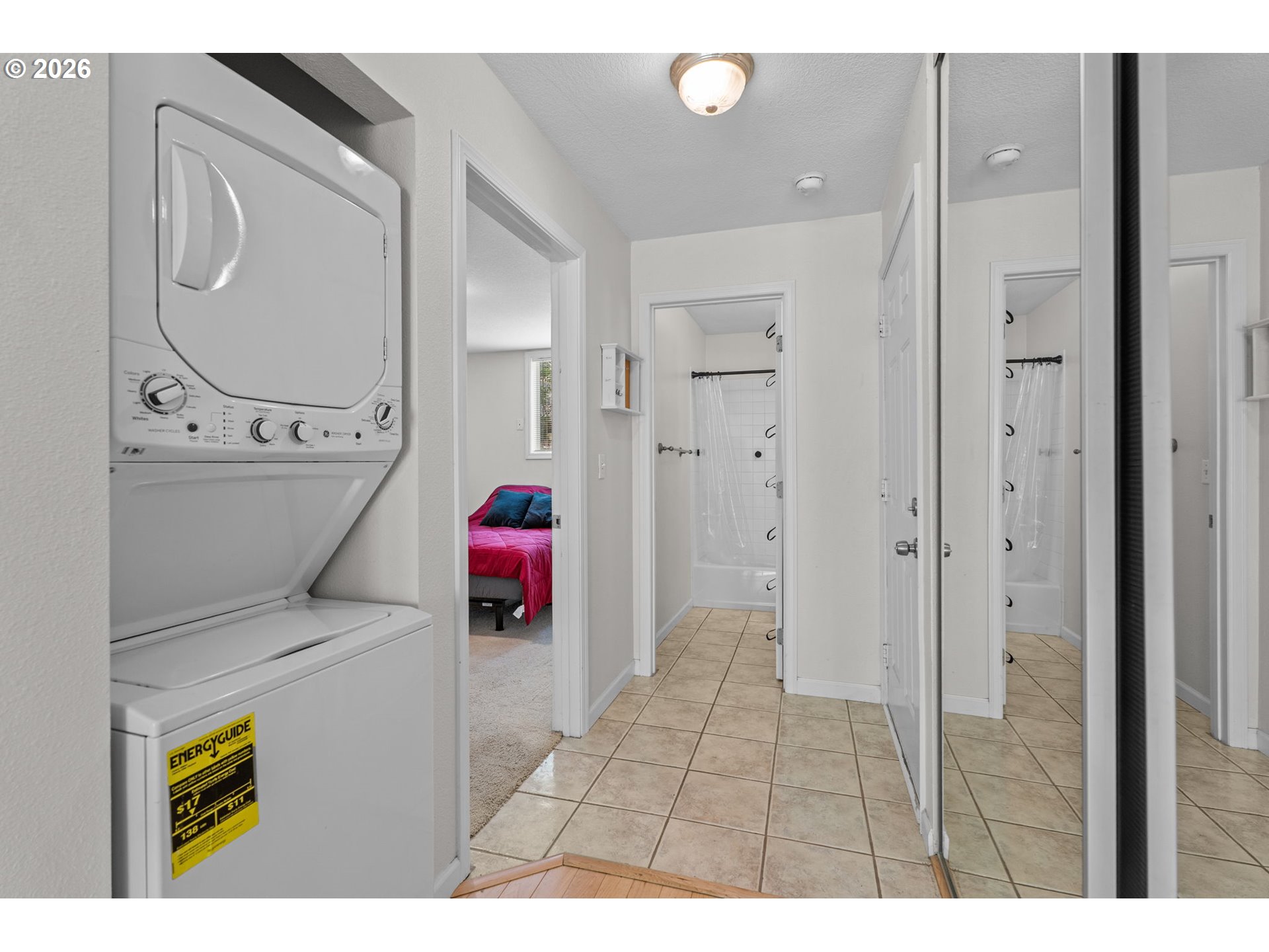 Tiled hallway with a washer and dryer, a bathroom doorway, and a bedroom with a bright pink bedspread.