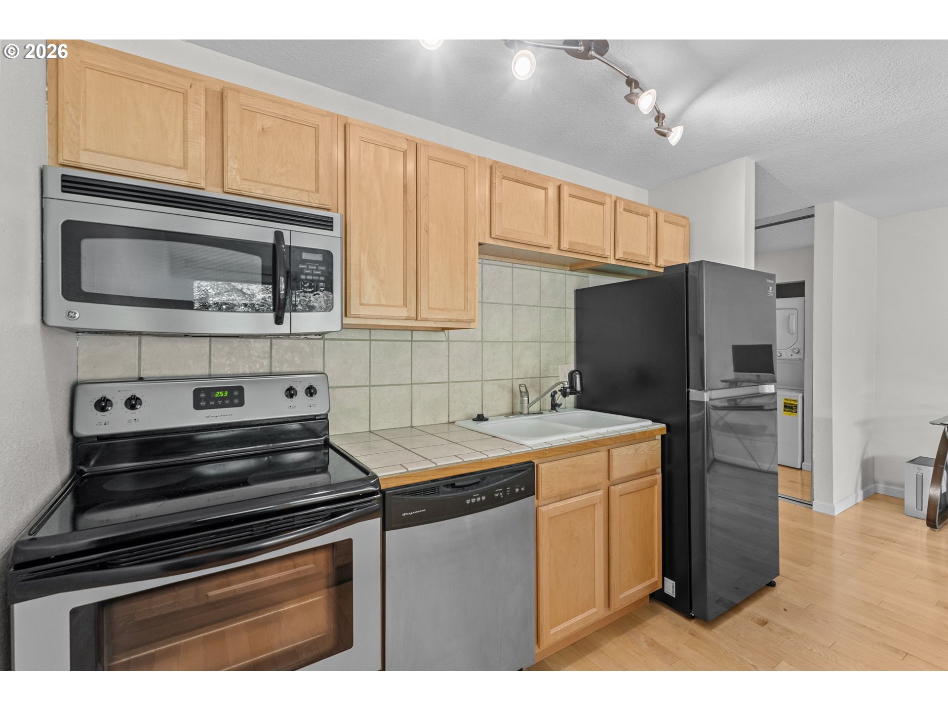Kitchen with light wood cabinets, tiled counters, and black and stainless steel appliances.