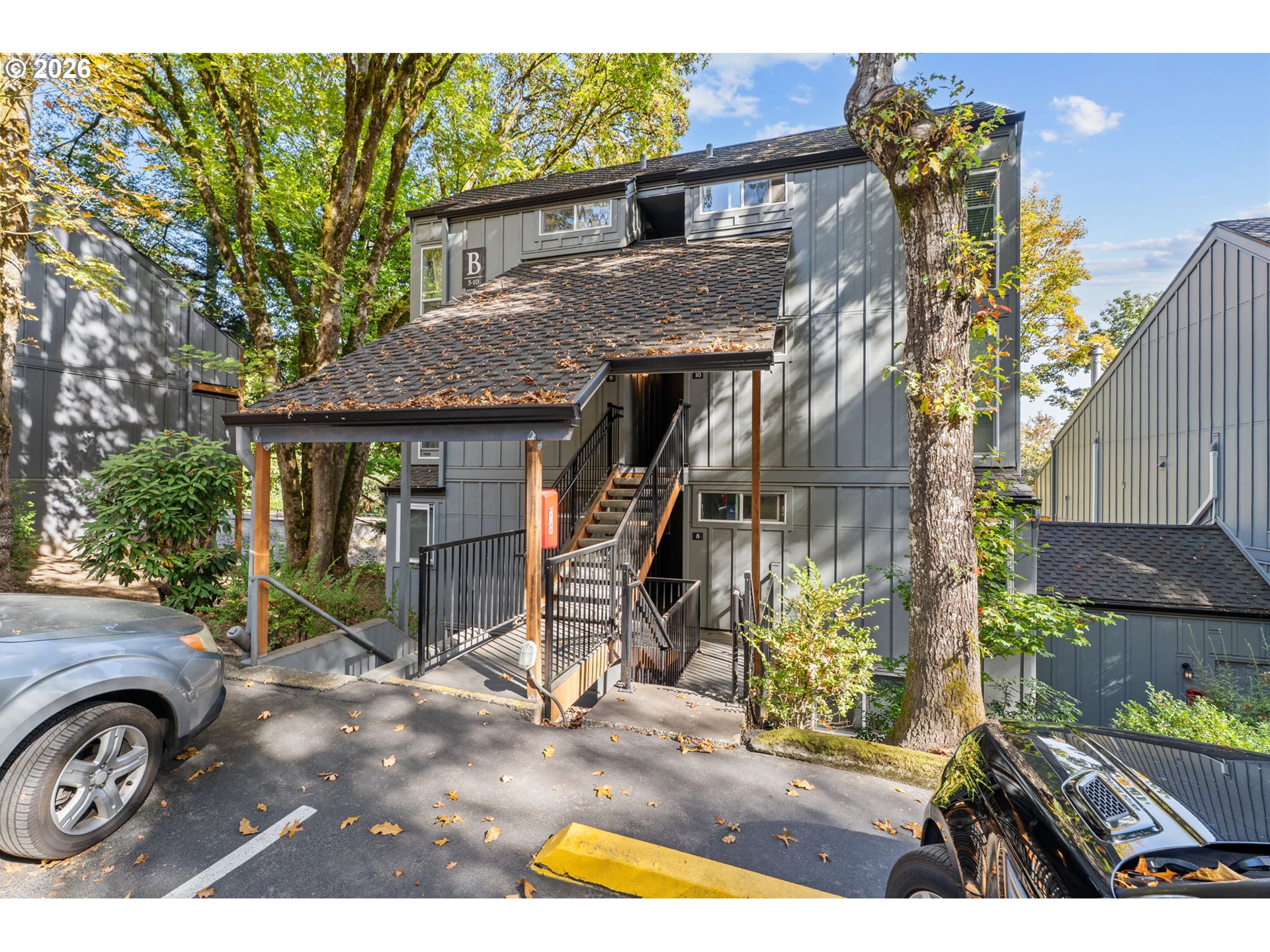 Gray multi-unit building with a covered entrance and exterior stairs beneath mature trees.