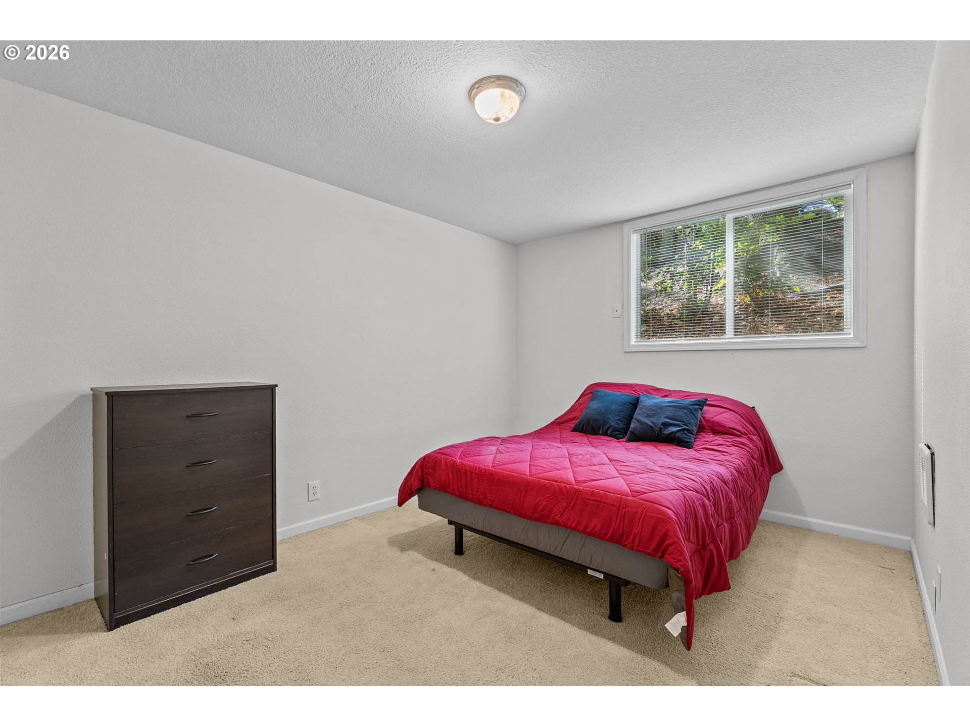 Bedroom with a red-quilted bed, dark dresser, beige carpet, and window overlooking greenery.