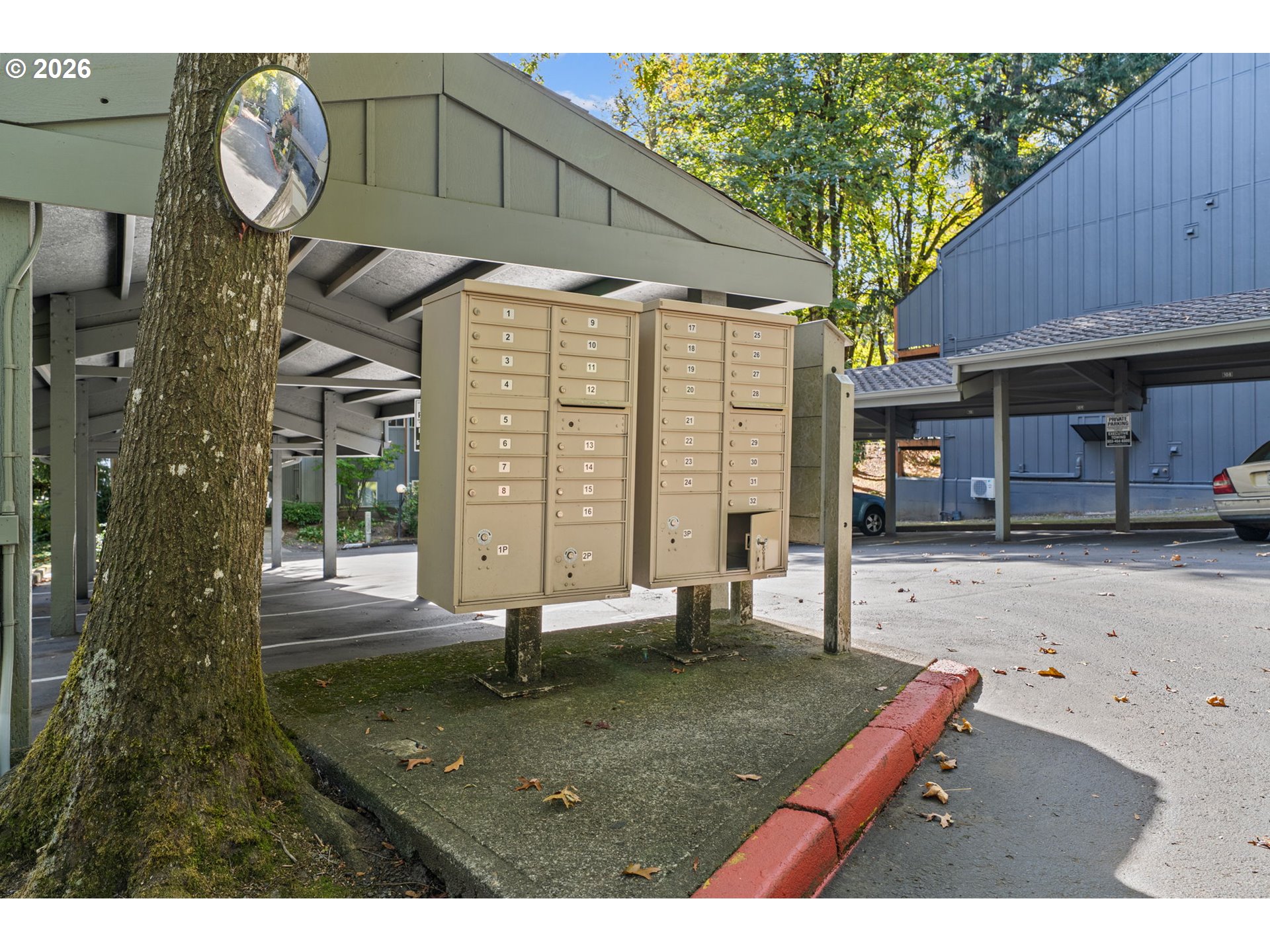 Numbered community mailboxes beneath a covered carport, beside a large tree and parking area.