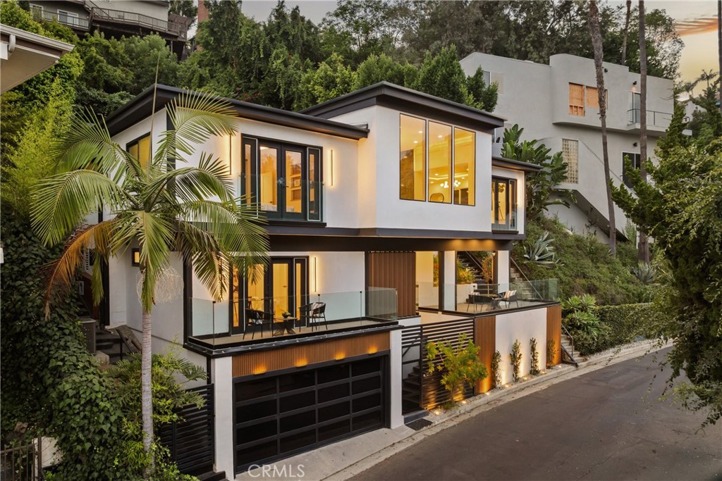 Modern white hillside home with glass balconies, tropical landscaping, and warmly lit windows at dusk.
