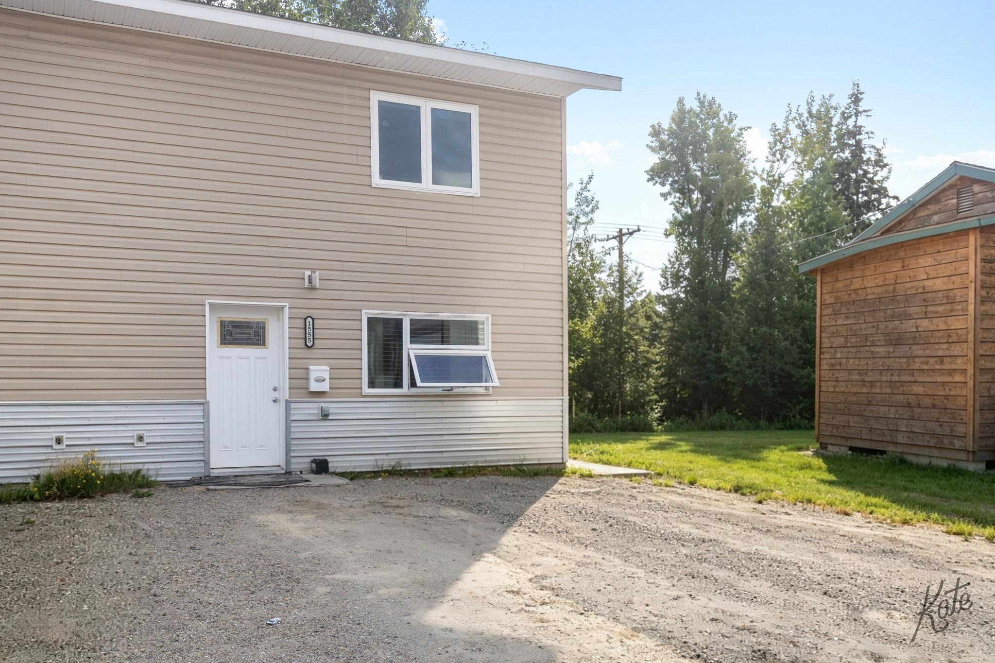 Two-story beige building beside a wooden outbuilding, with a gravel driveway and grassy yard.