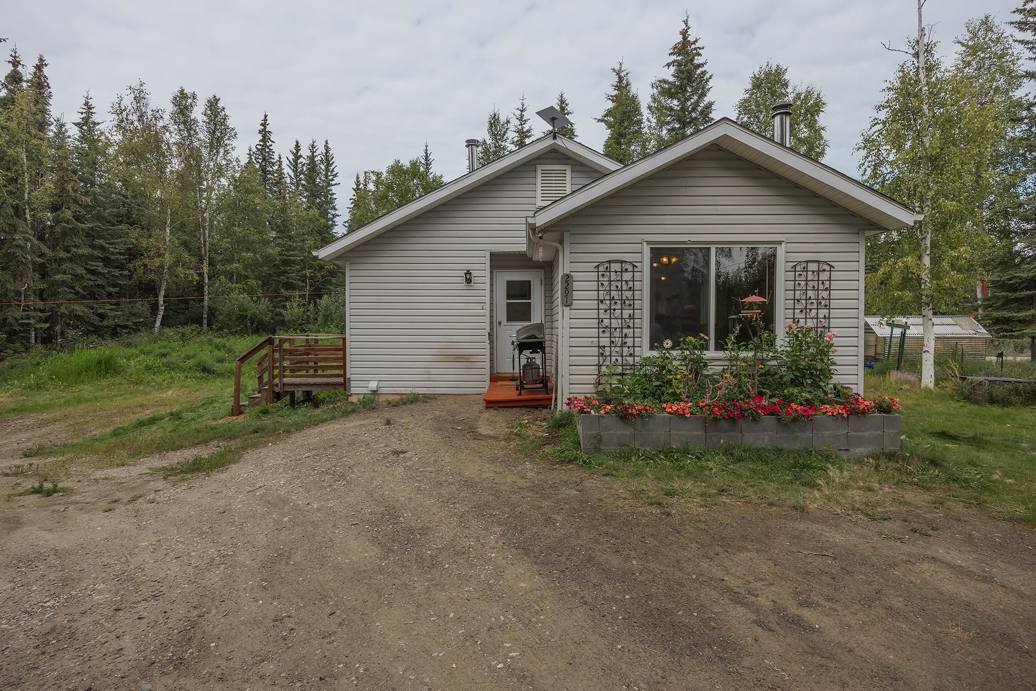 Gray-sided house with a gravel driveway, flower bed, and wooded surroundings.
