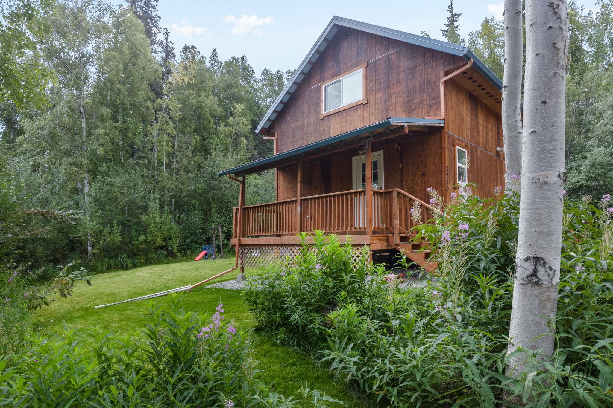 Wood-sided two-story home with a covered porch, garden flowers, and trees around the lawn.