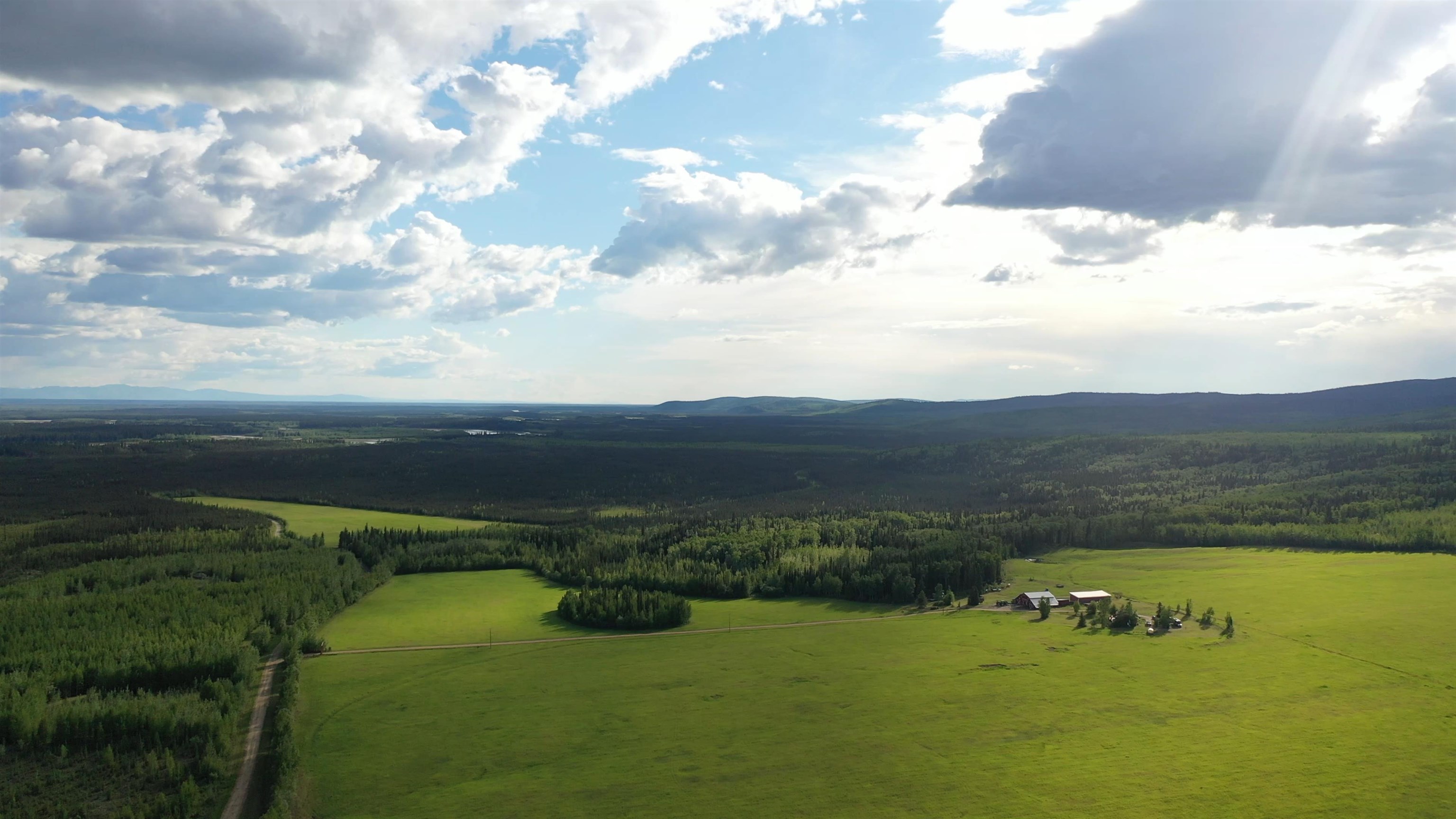 Aerial view of broad green fields, forest, farm buildings, and distant hills.