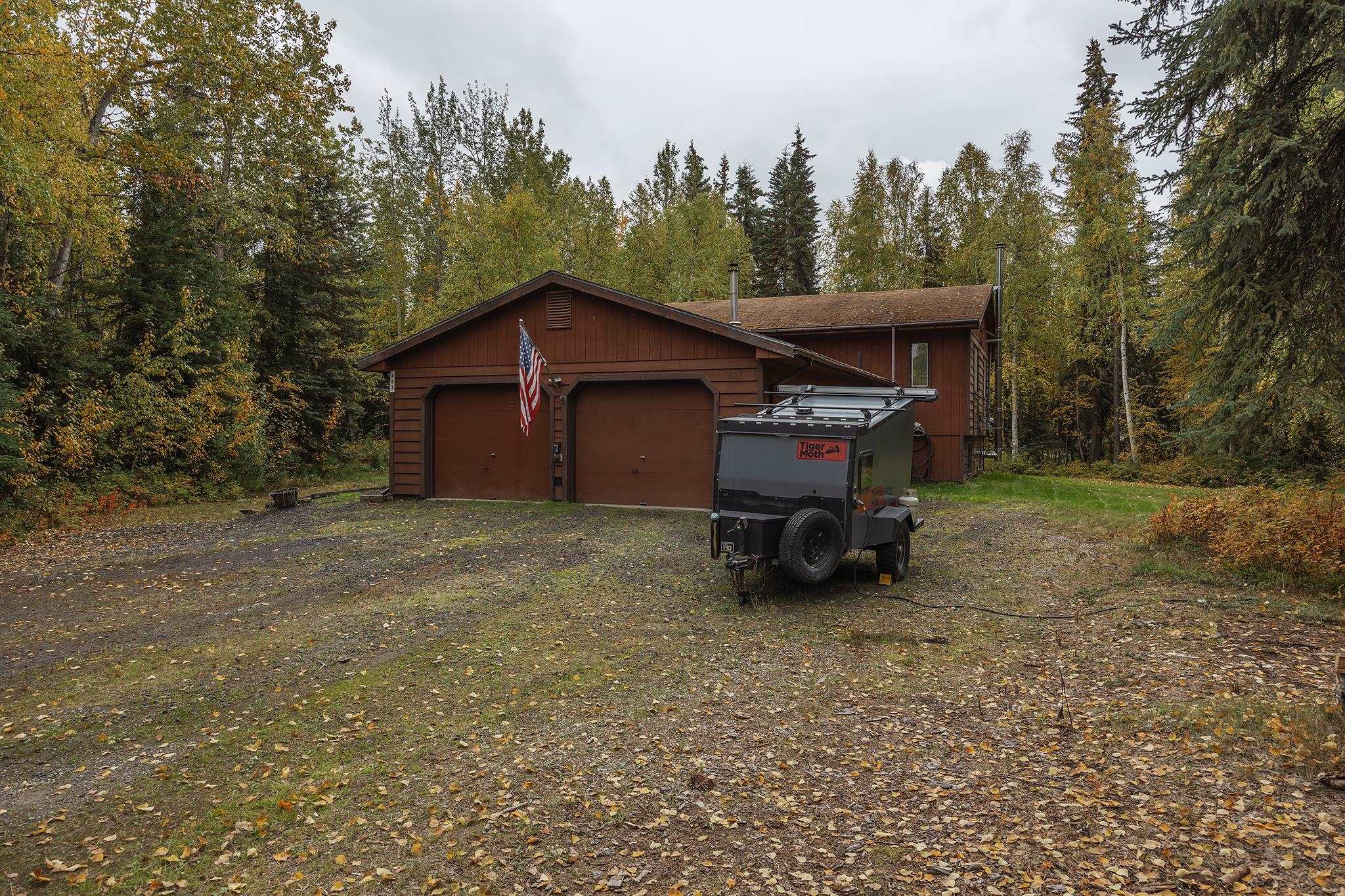 Brown two-story house with a double garage, wooded surroundings, and a utility trailer in the driveway.