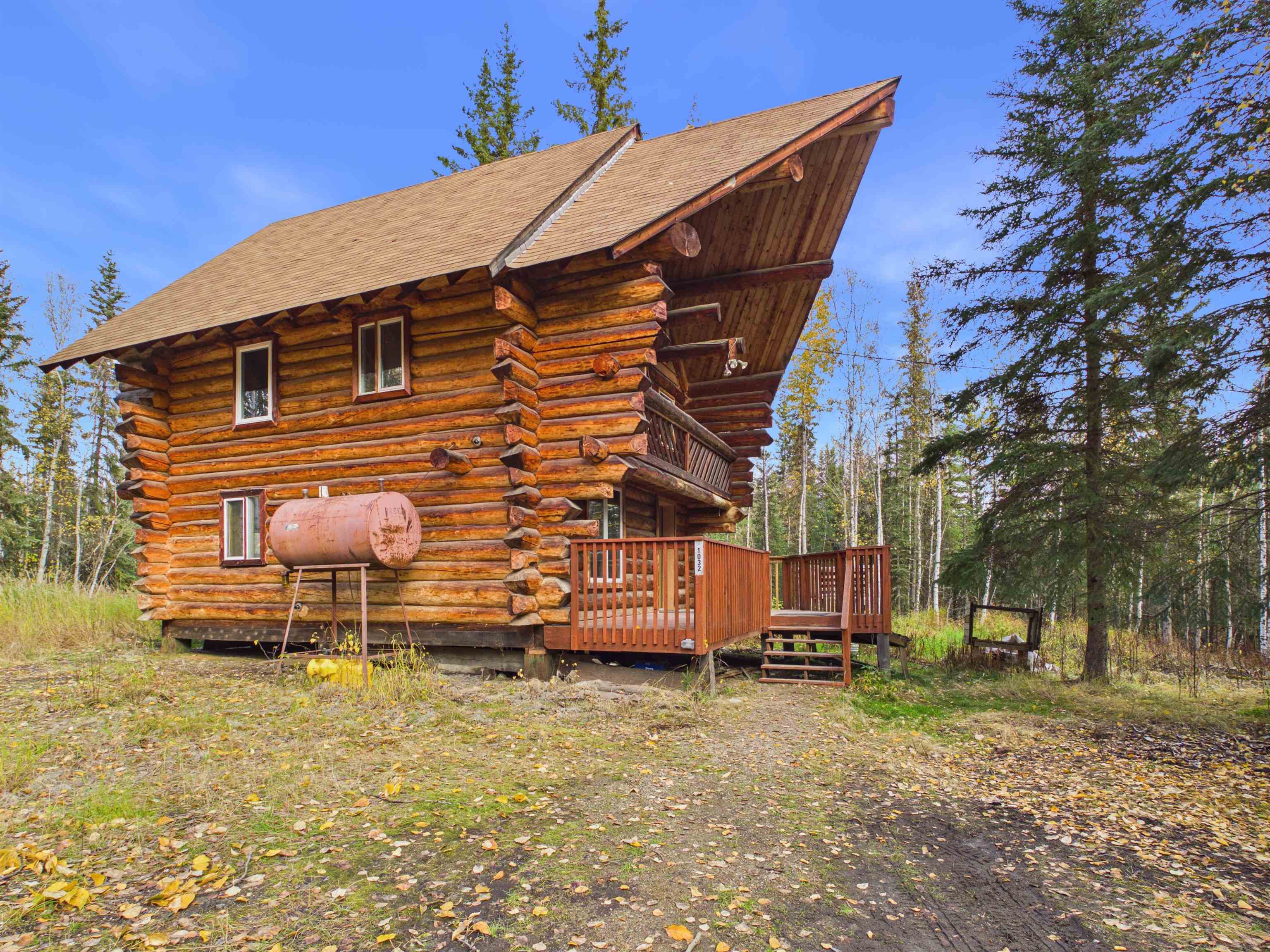 Two-story log cabin with a steep roof, raised porch and balcony among autumn trees.