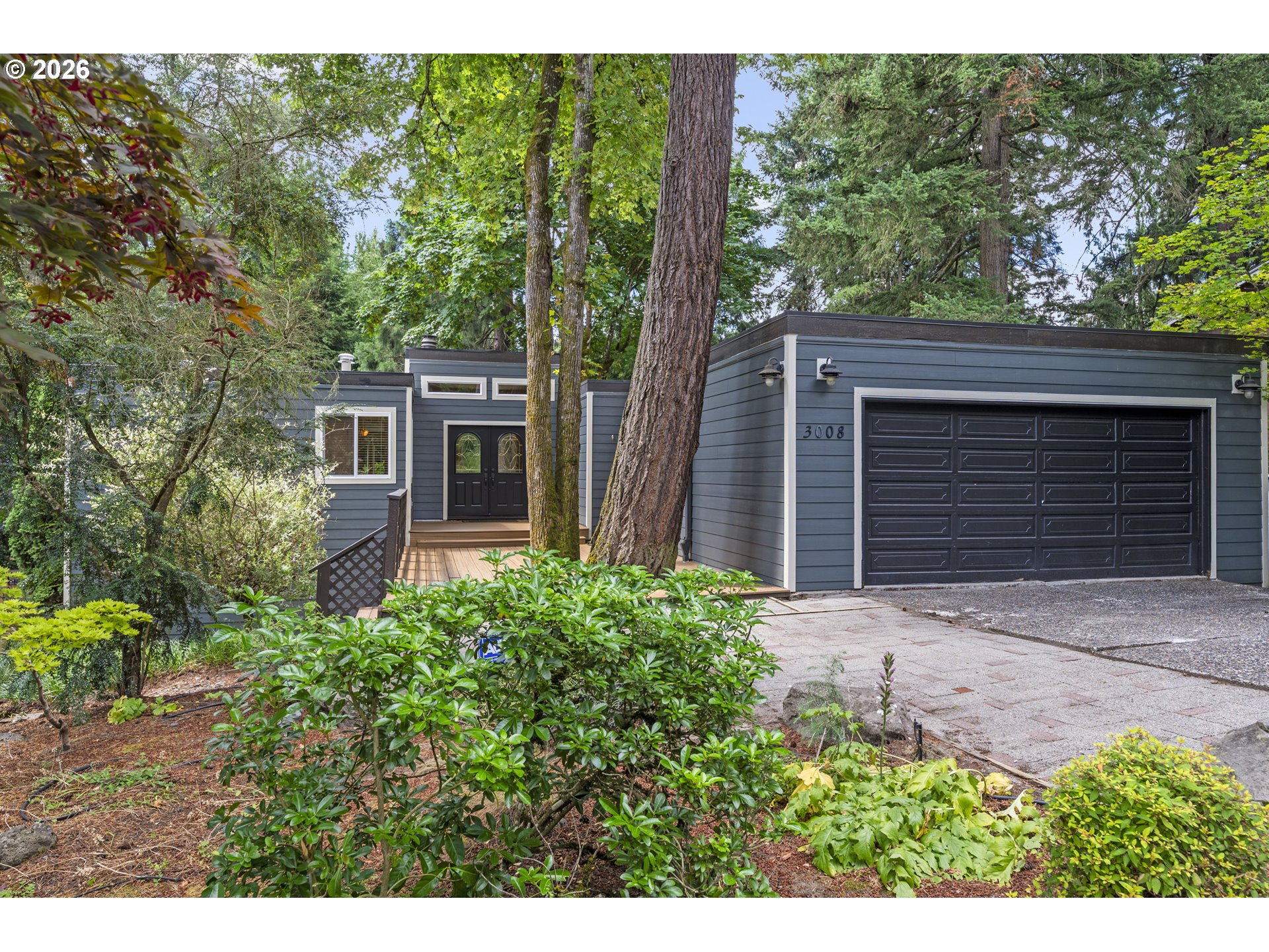 Gray home with a black double garage door, front deck, and mature trees.