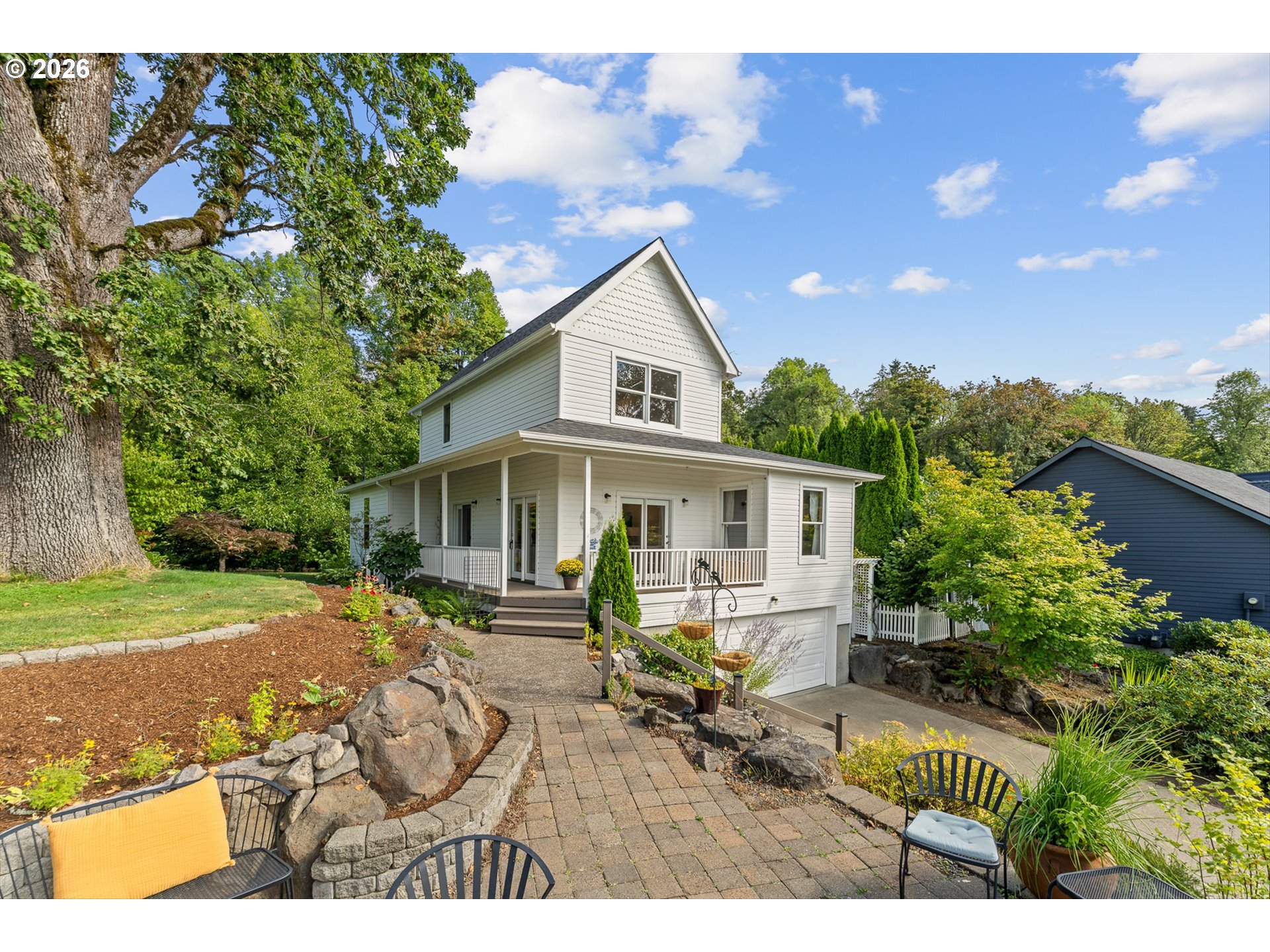 White two-story house with a covered porch, landscaped garden, and paved patio with outdoor seating.