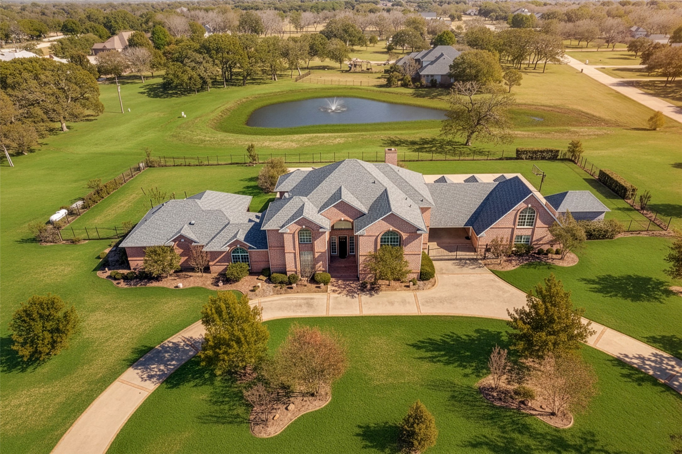 Aerial view of a large brick home with a curved driveway and fenced lawn beside a pond with a fountain.