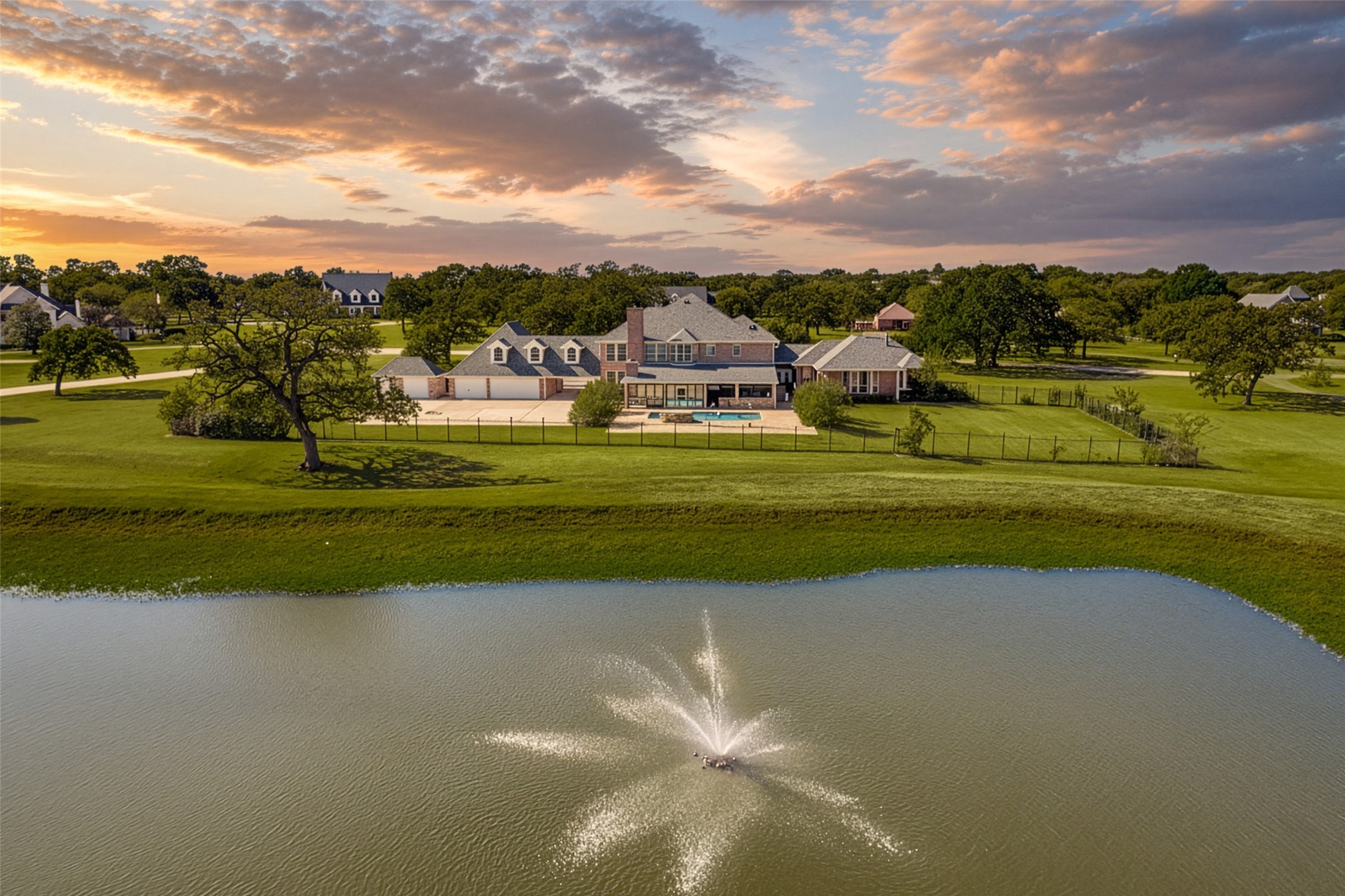 Aerial view of a large home beside a pond with a fountain, pool, and broad lawn.