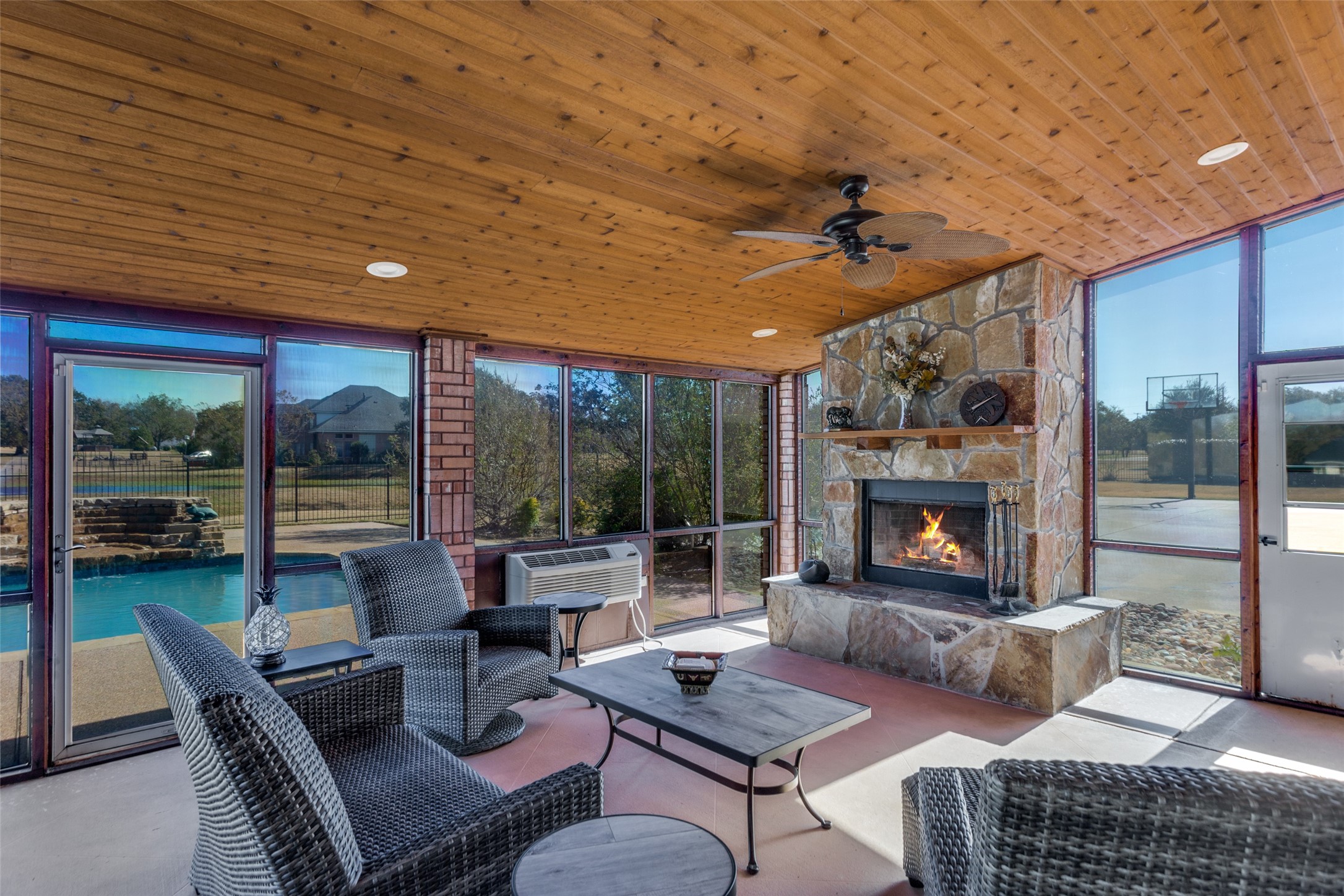Enclosed patio with wicker seating, a stone fireplace, and views of the pool through large windows.