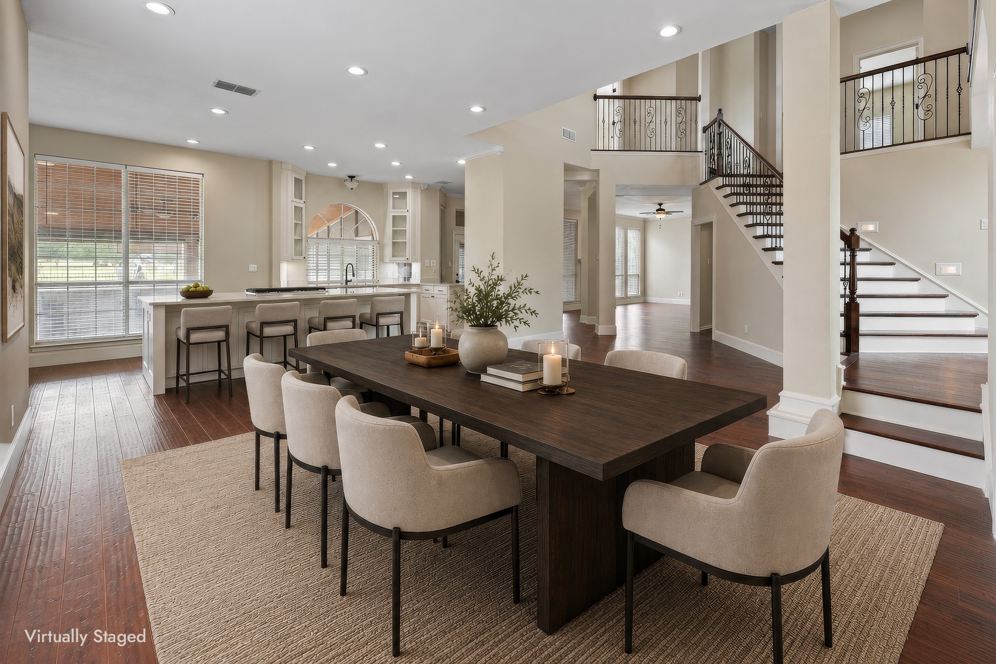 Open-plan dining room with a long dark table, upholstered chairs, kitchen breakfast bar, and decorative staircase.