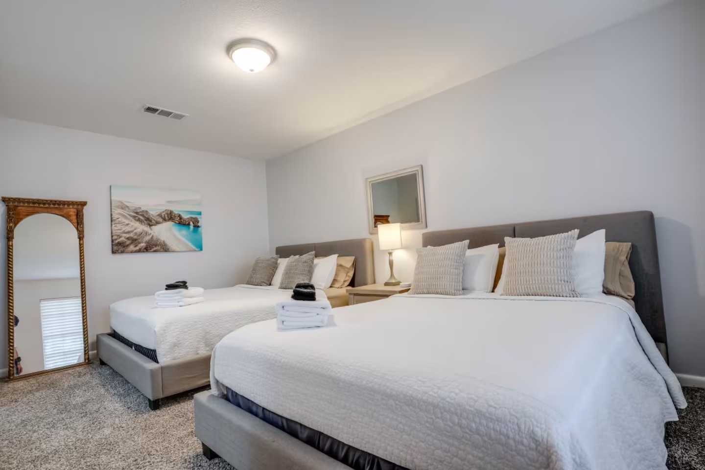 Bedroom with two white-dressed beds, gray upholstered headboards, and a lamp between them.