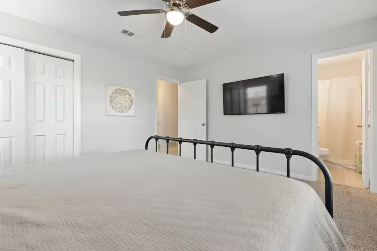 Bedroom with a black metal bed frame, ceiling fan, wall-mounted TV, and bathroom doorway.