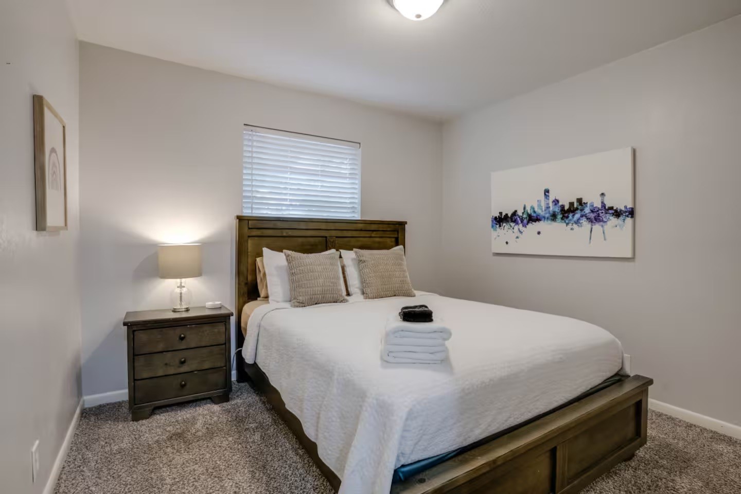 Bedroom with a dark wood bed, white bedding, gray carpet, and colorful cityscape wall art.
