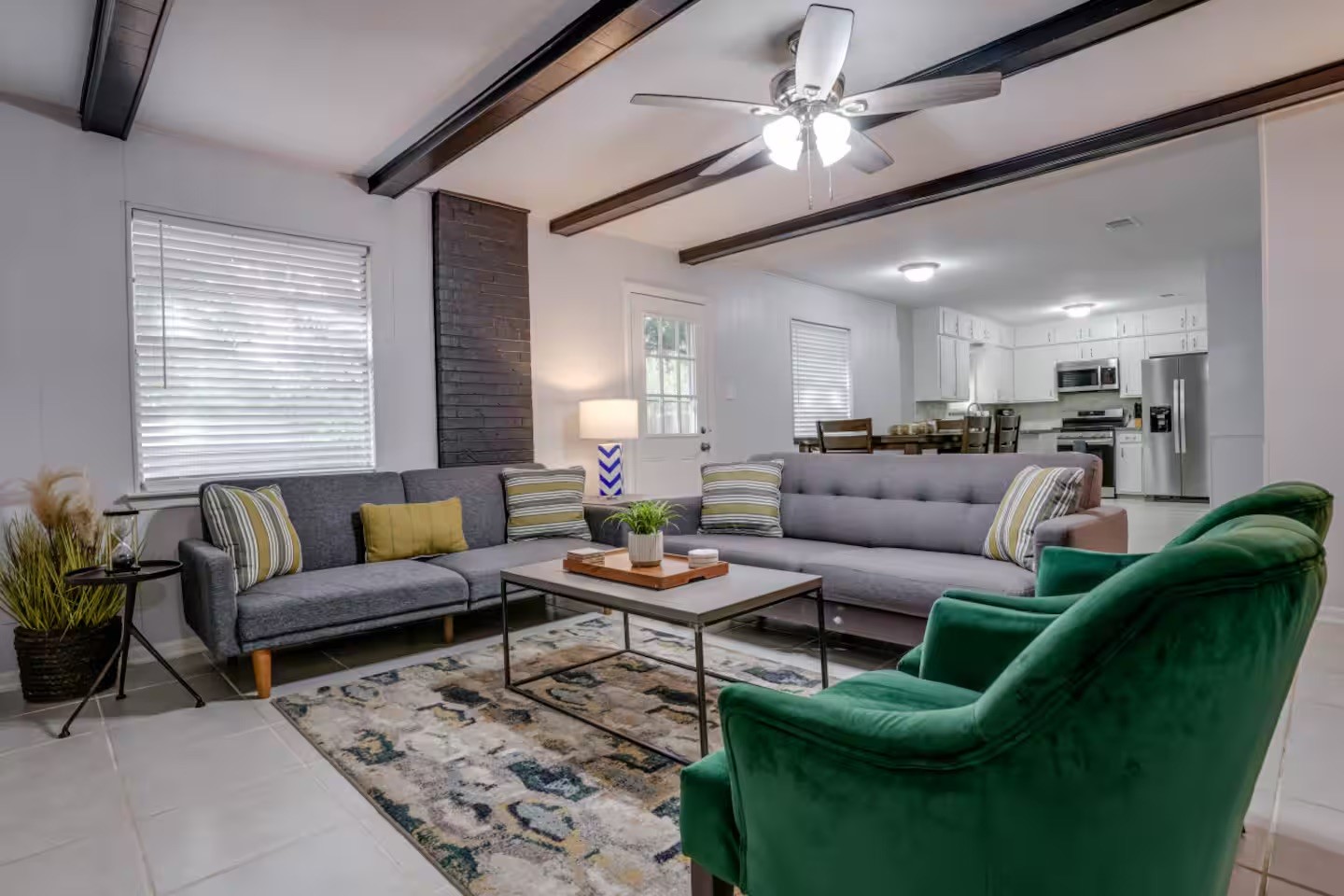Living room with a gray sectional, green armchairs, exposed ceiling beams, and an open kitchen.