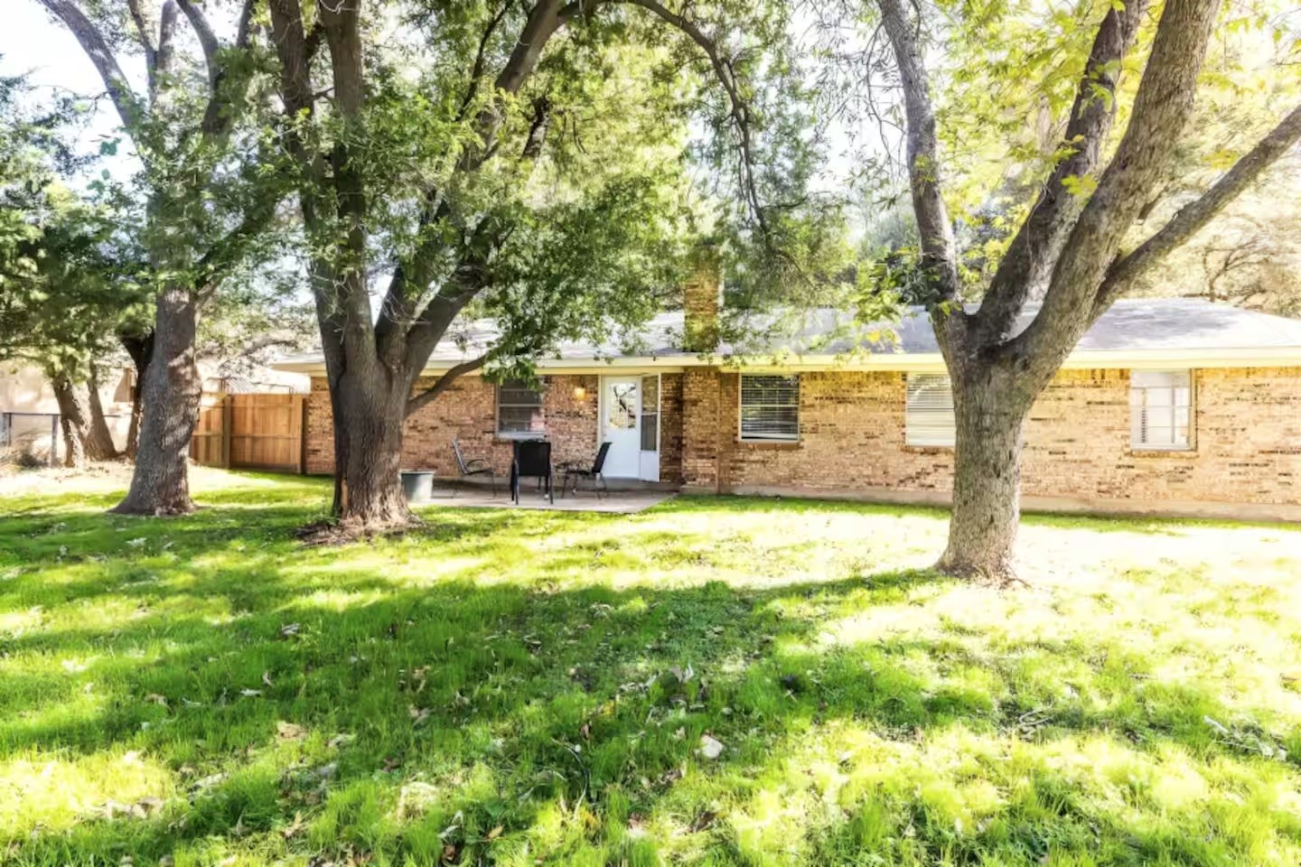 Single-story brick home with a small patio, shaded lawn, and mature trees.