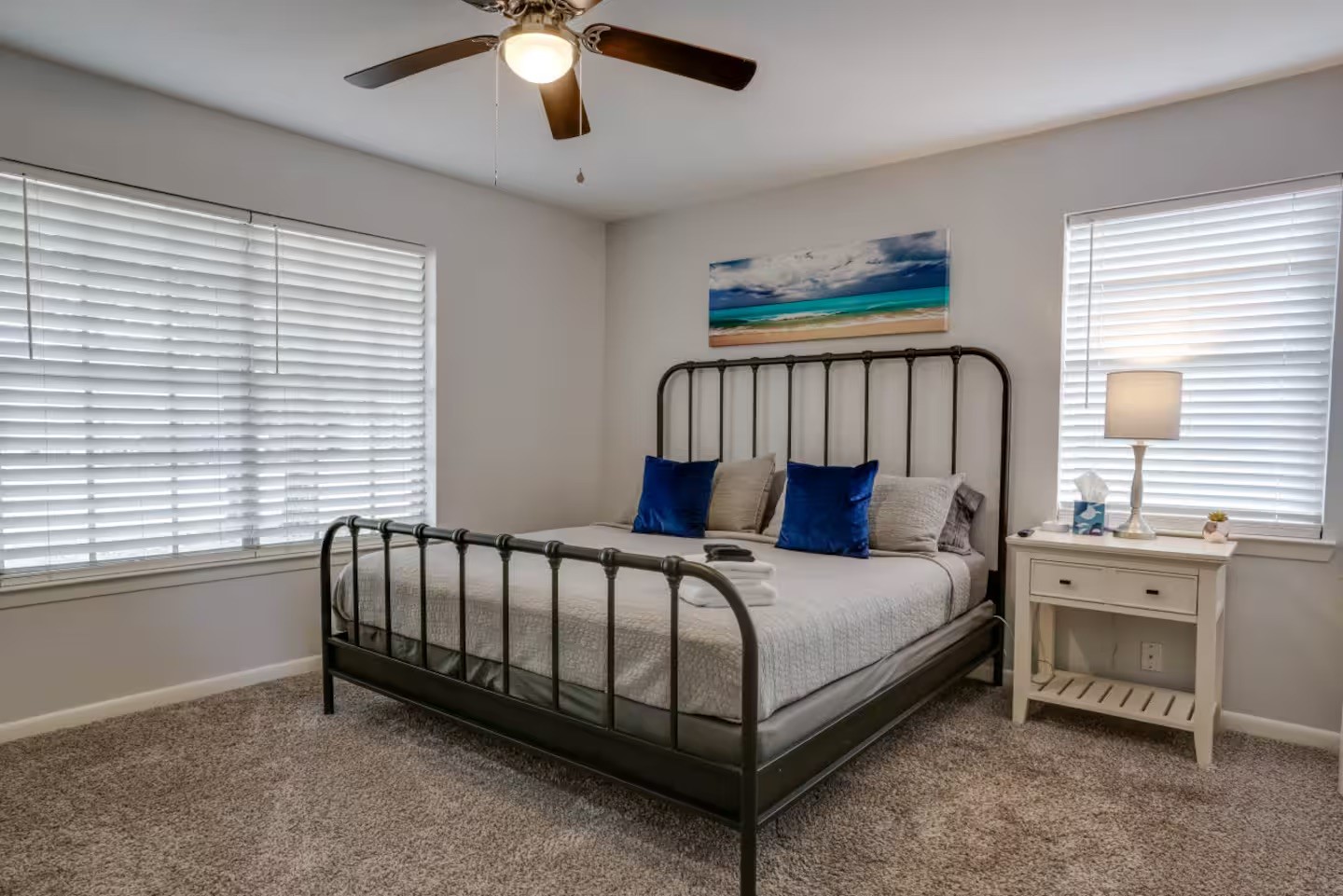 Furnished bedroom with a dark metal bed frame, blue accent pillows, wide windows and a ceiling fan.