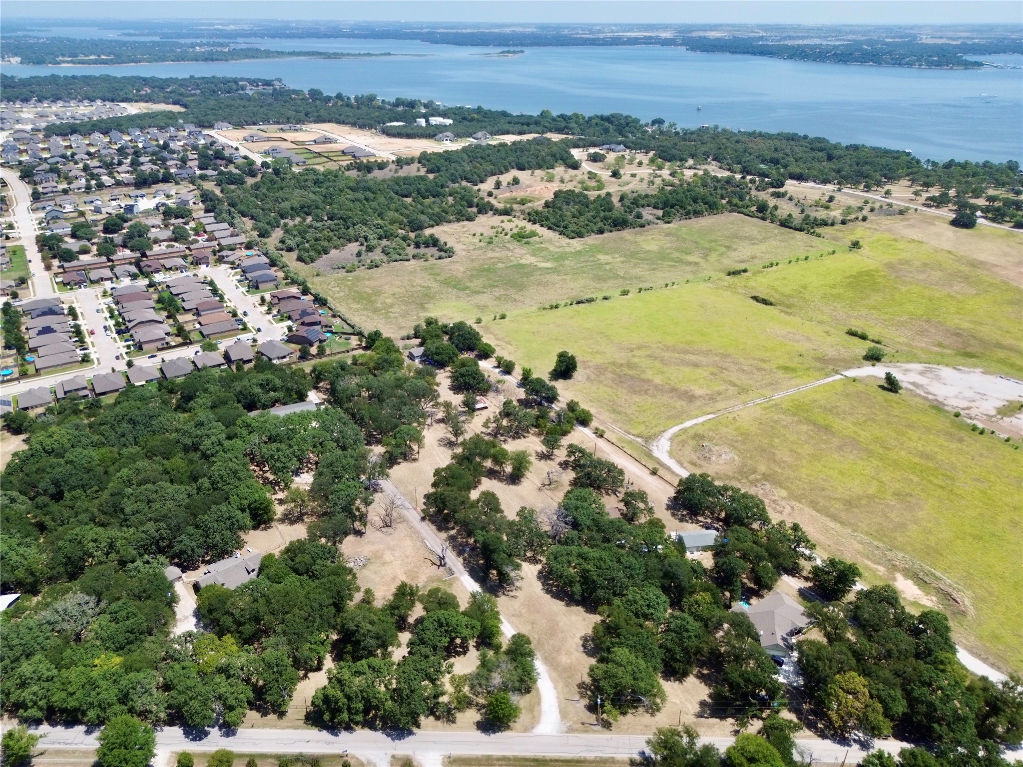 Aerial view of open fields and mature trees beside a neighborhood, with a lake in the background.
