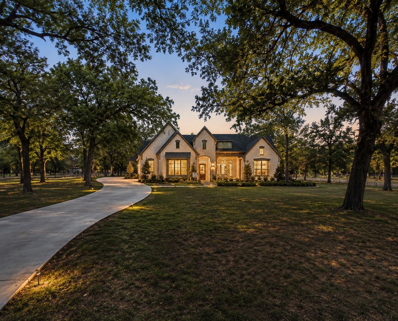 Gabled house with glowing windows at dusk, framed by mature trees and a curved driveway.