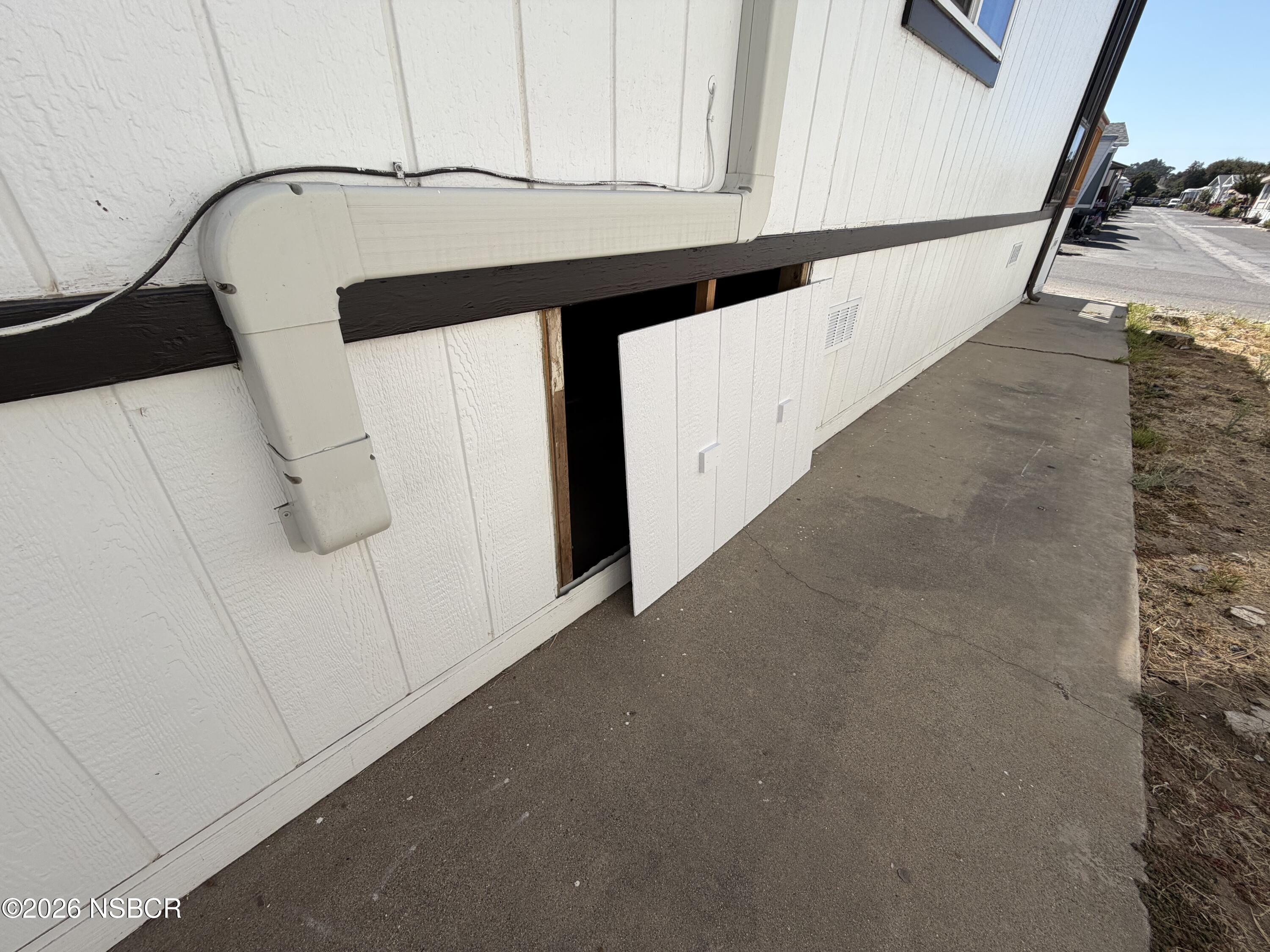 White-sided exterior wall with an open lower access panel beside a concrete walkway.