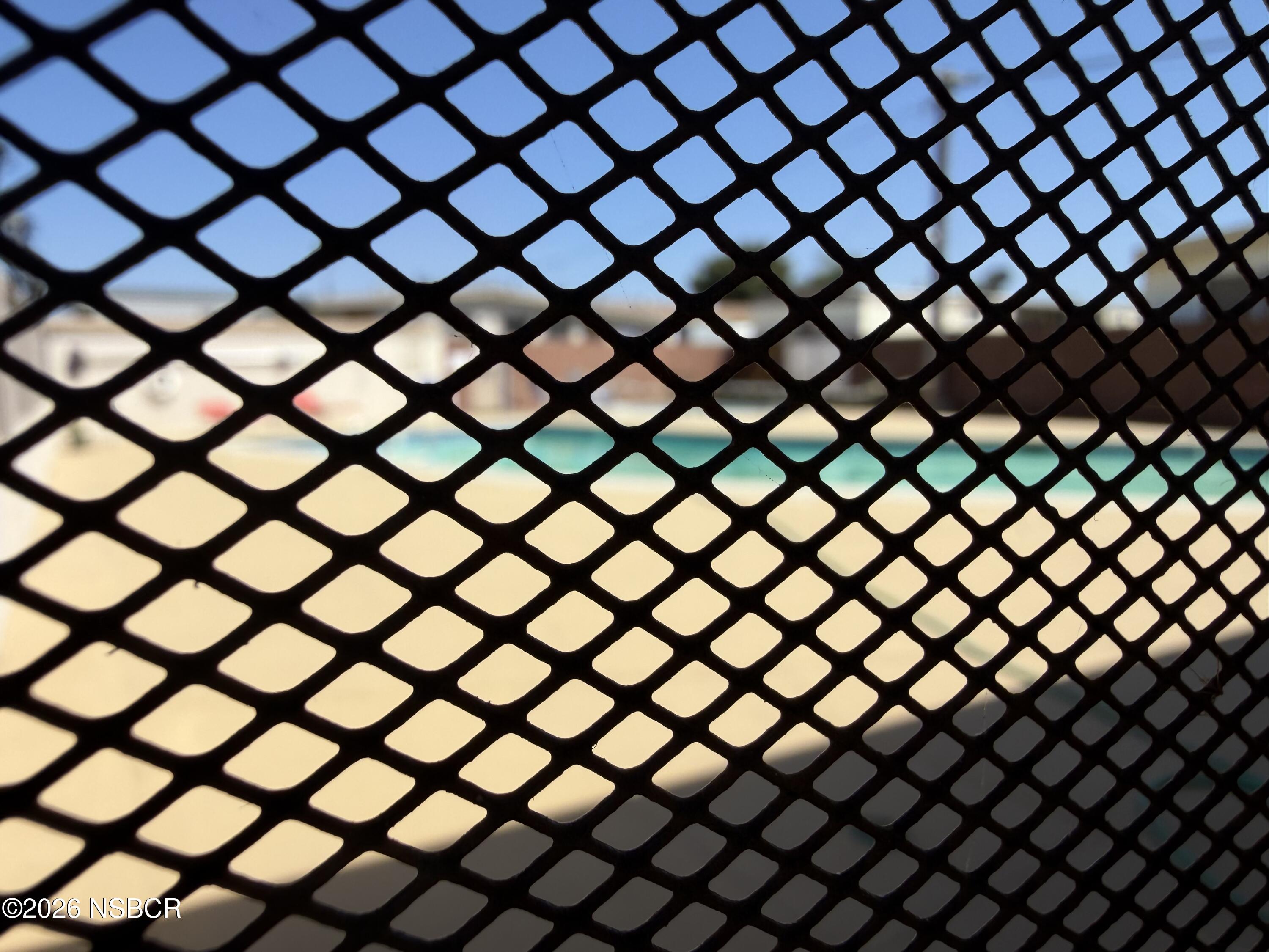 Swimming pool seen through a black mesh fence, with turquoise water and a pale deck.