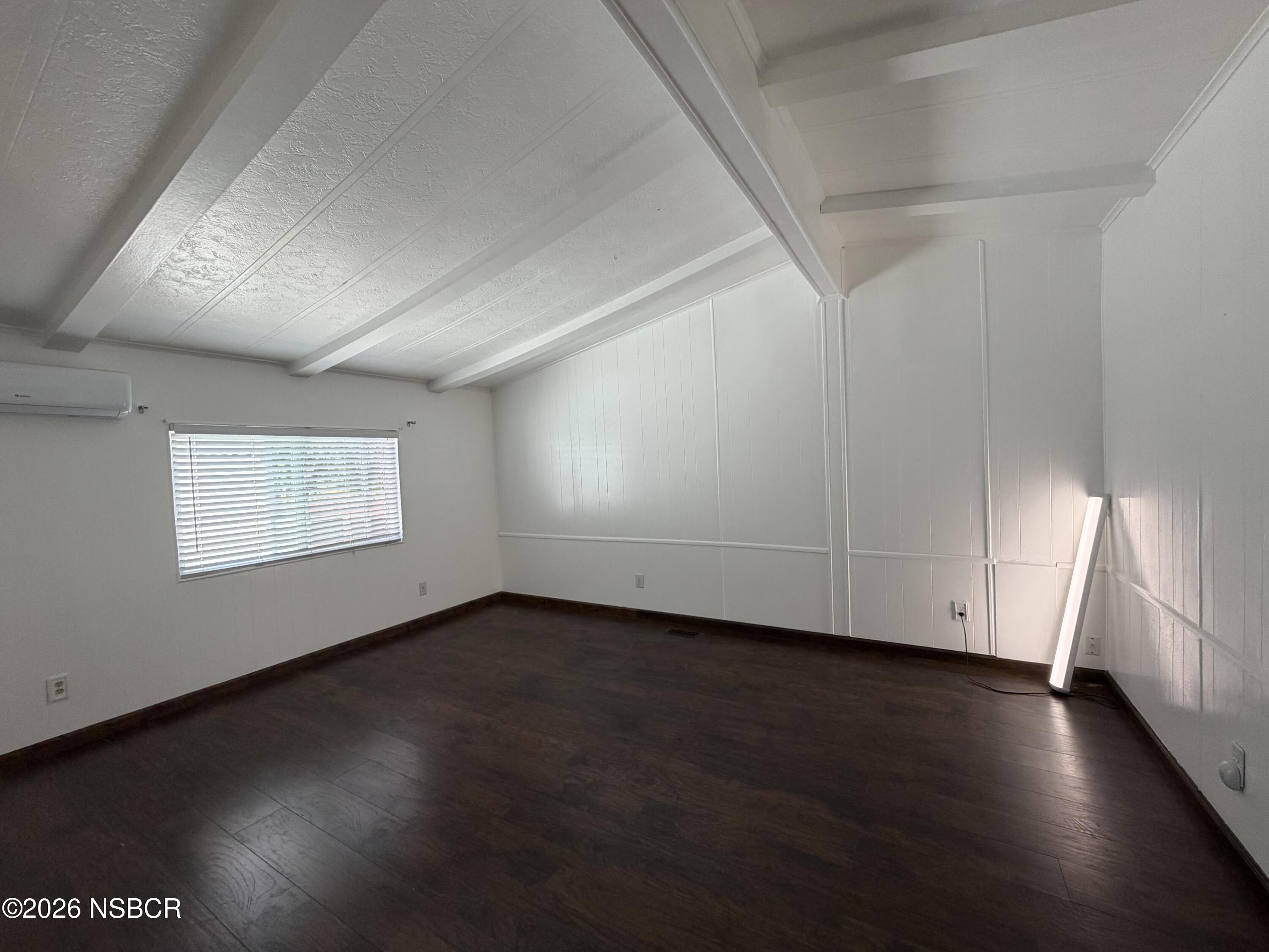 Empty room with dark wood flooring, a white beamed ceiling, window blinds, and a wall-mounted air conditioner.