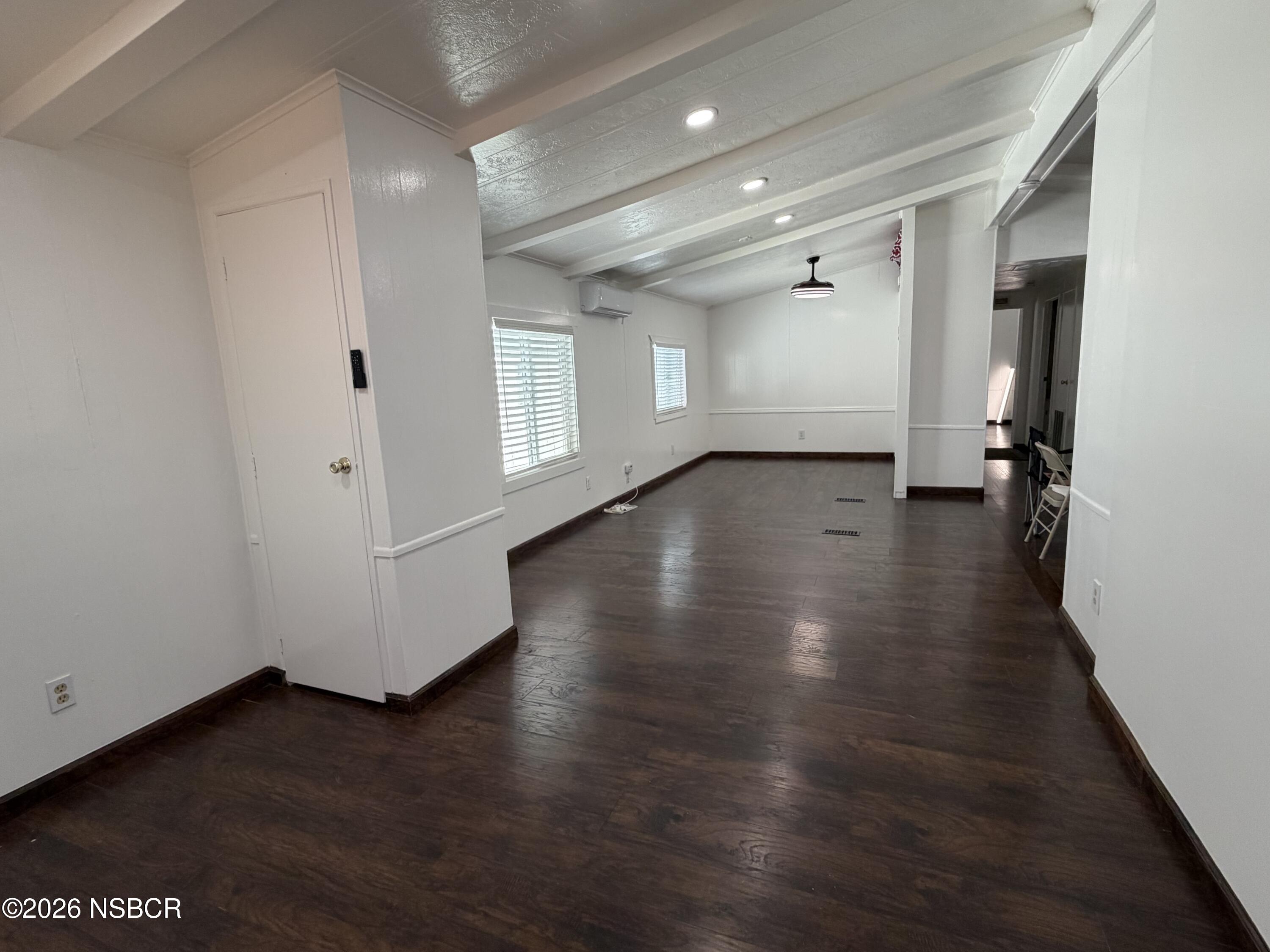 Vacant living area with dark flooring, white walls, exposed ceiling beams, and windows with blinds.