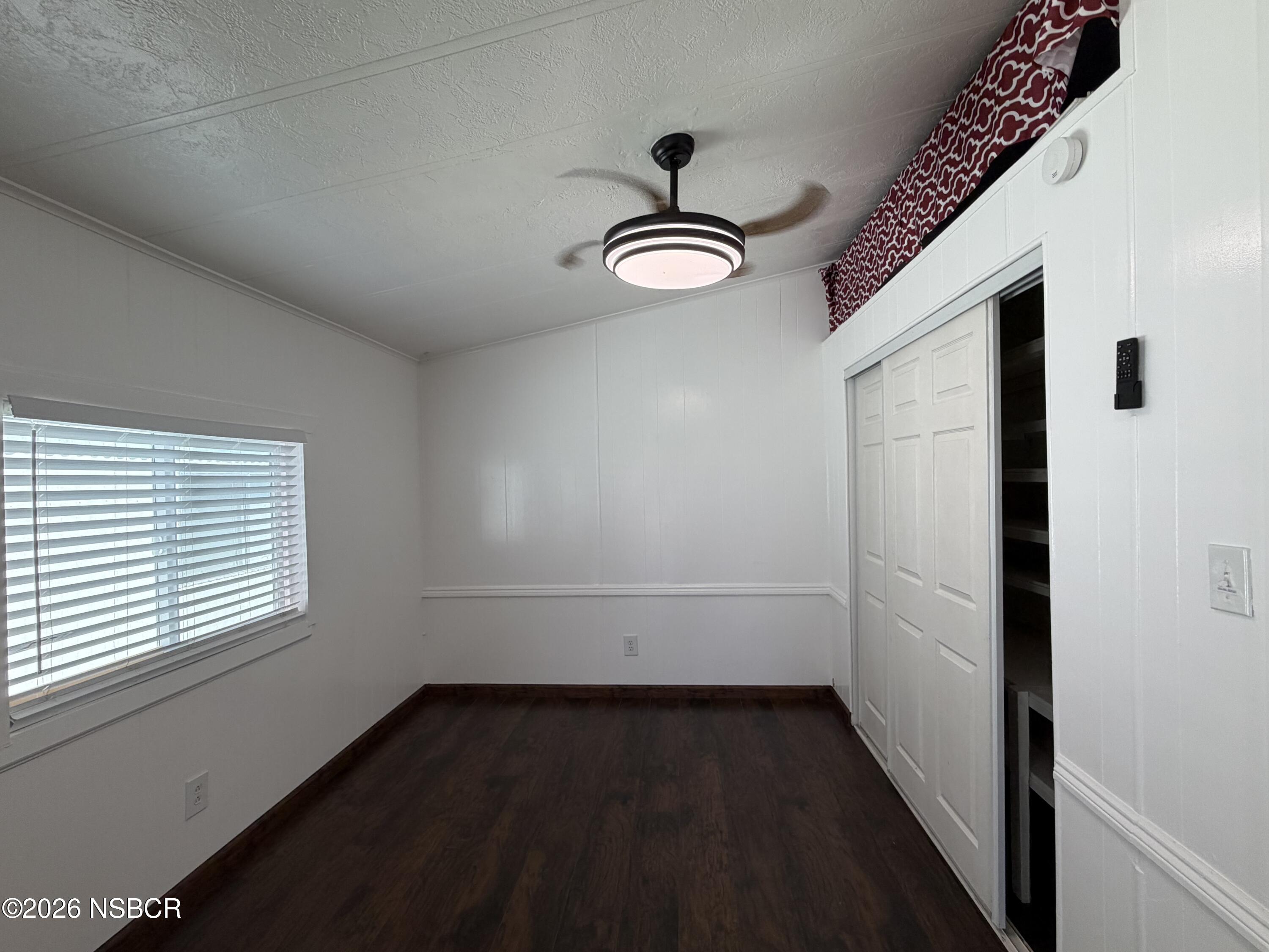 Empty bedroom with dark wood floors, window blinds, sliding closet doors, and a ceiling fan with light.