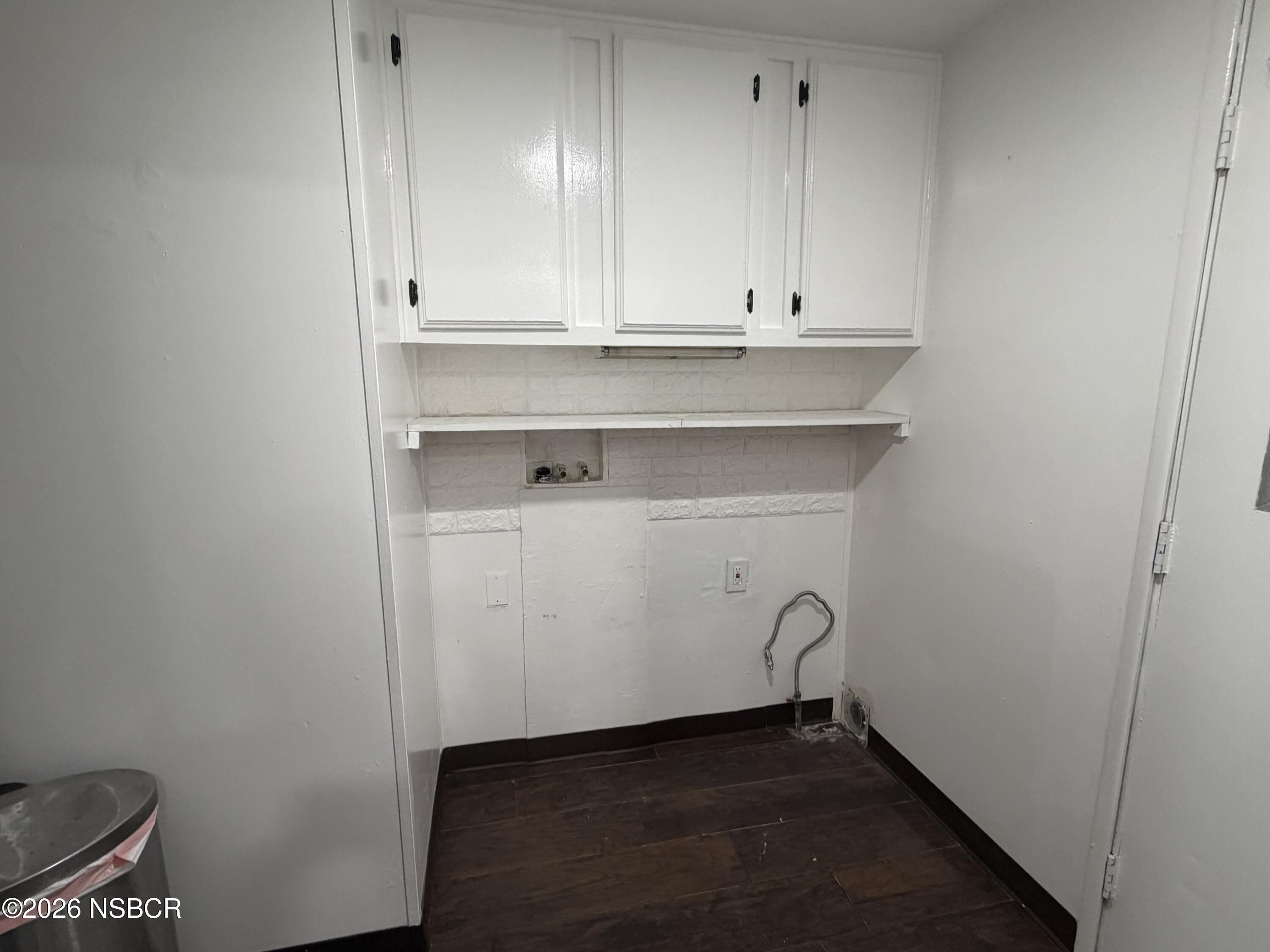 Vacant laundry area with white cabinets, a wall shelf, exposed washer hookups, and dark flooring.