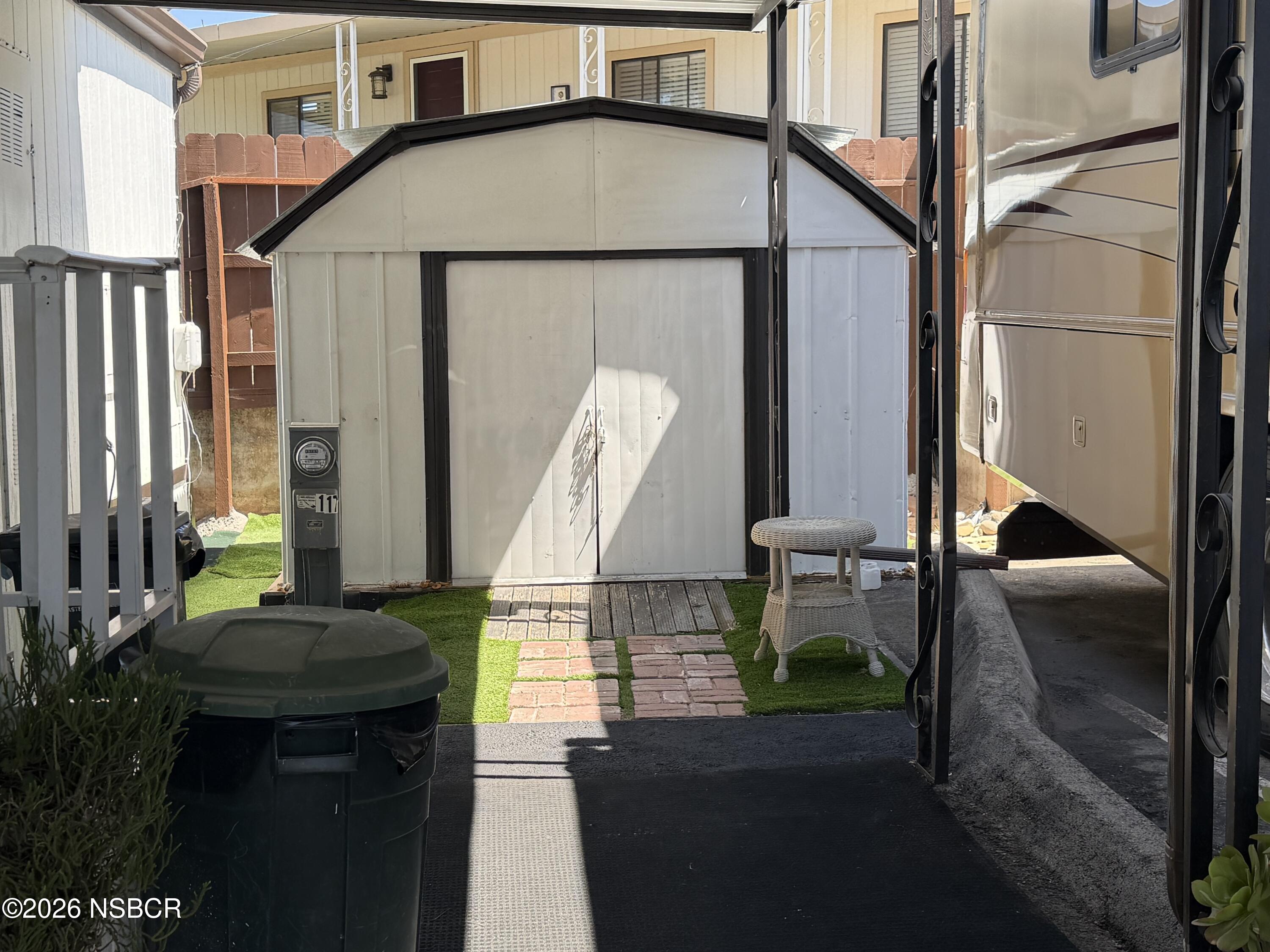 Covered outdoor area with a white storage shed, artificial turf, brick stepping stones, and a parked recreational vehicle.