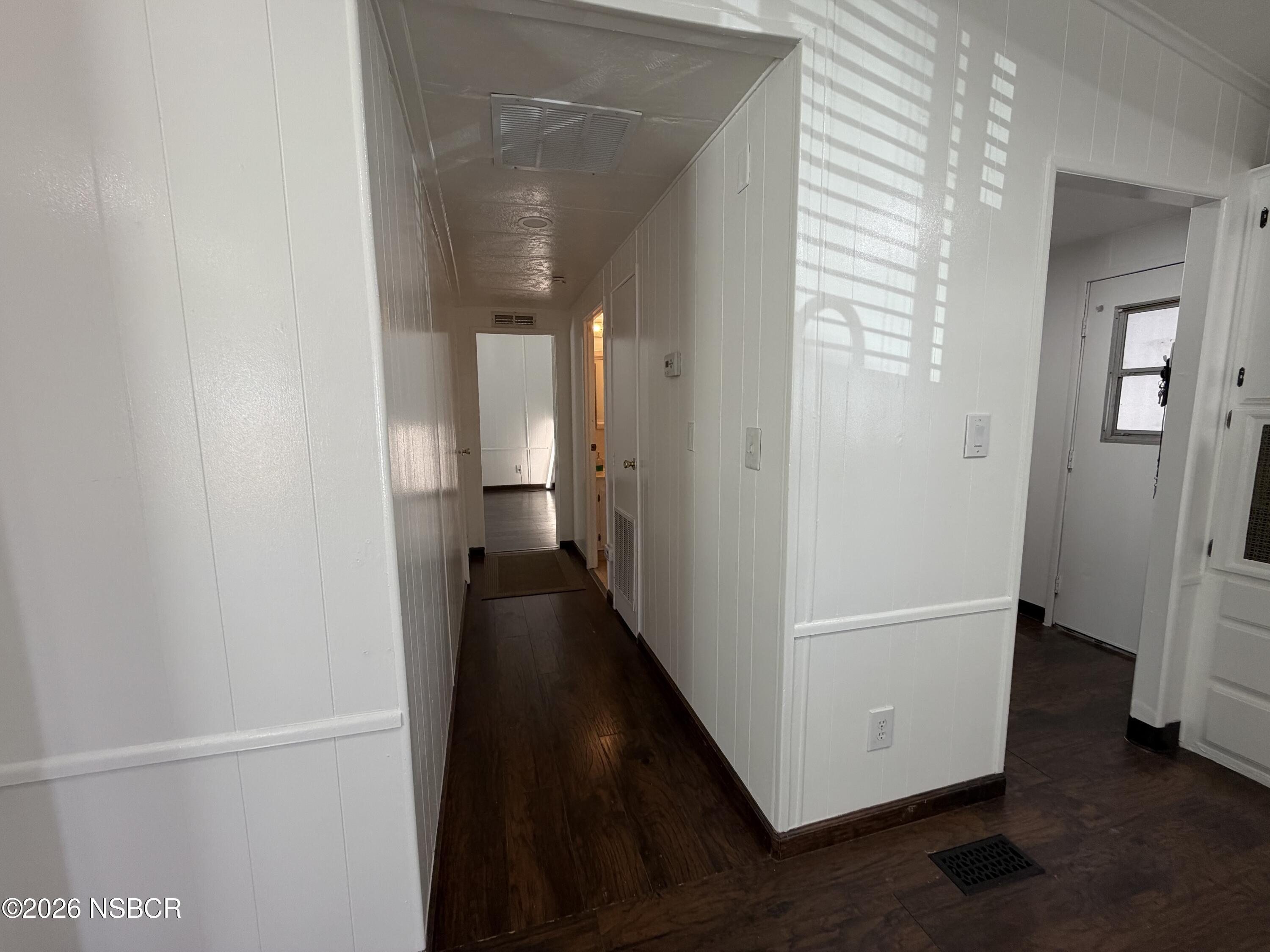 Hallway with dark wood floors, white paneled walls, and a side entry door.
