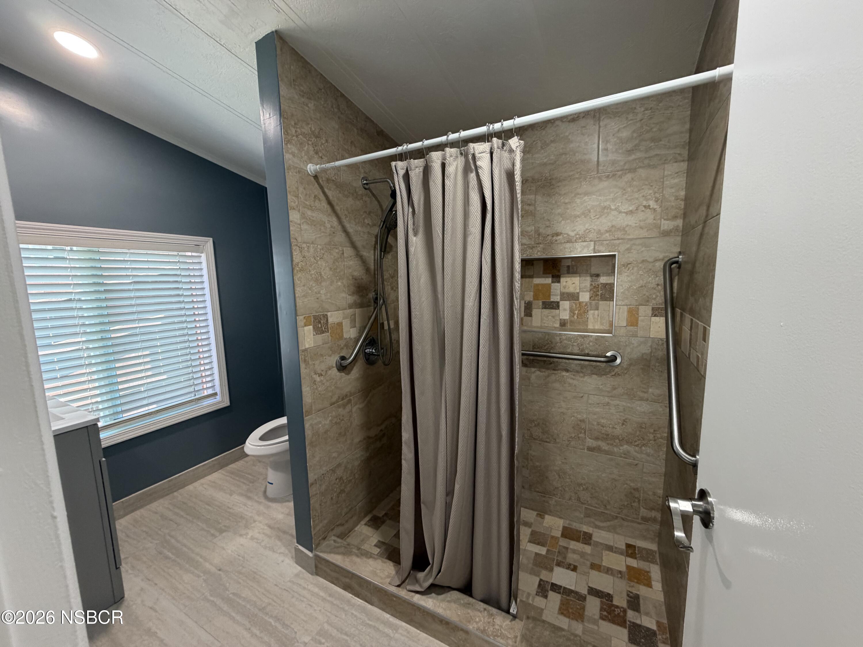 Bathroom with tiled walk-in shower, mosaic floor, grab bars, and a window with blinds.