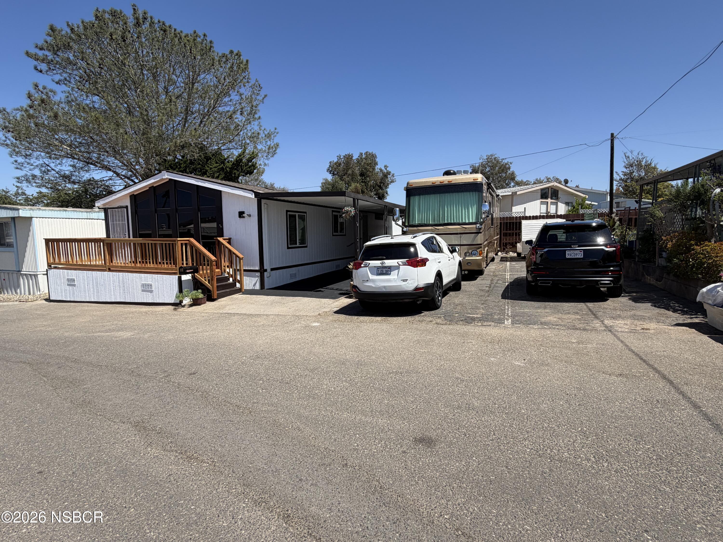 Manufactured home with a wooden porch, next to parked vehicles in a residential community.
