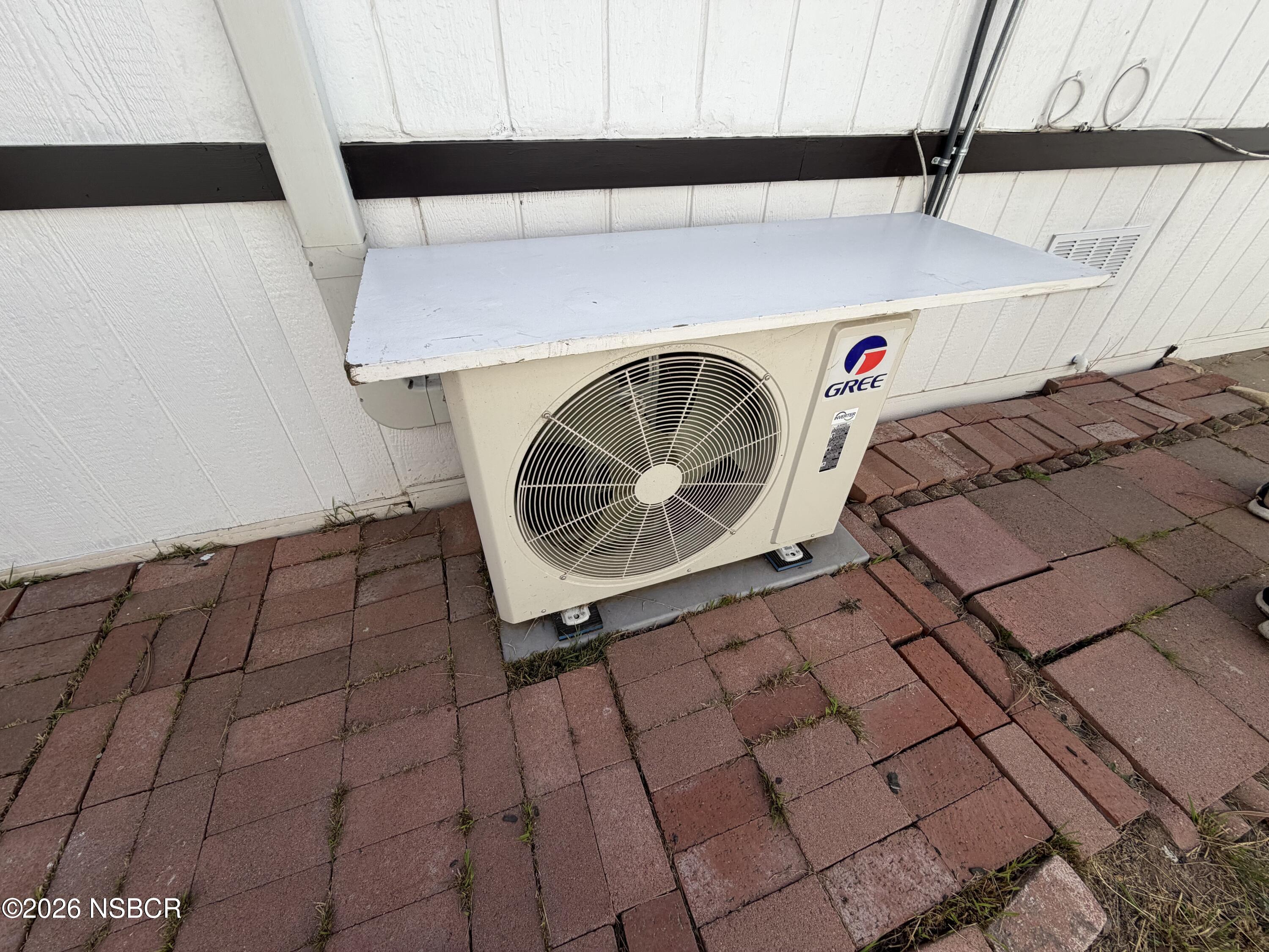Outdoor GREE heat pump condenser beneath a protective cover, beside a white-sided building and brick pavers.