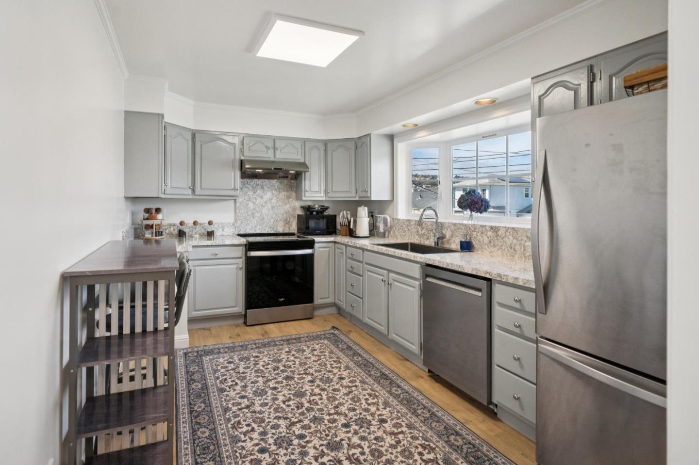 Kitchen with gray cabinets, stainless steel appliances, a window above the sink, and a patterned rug.