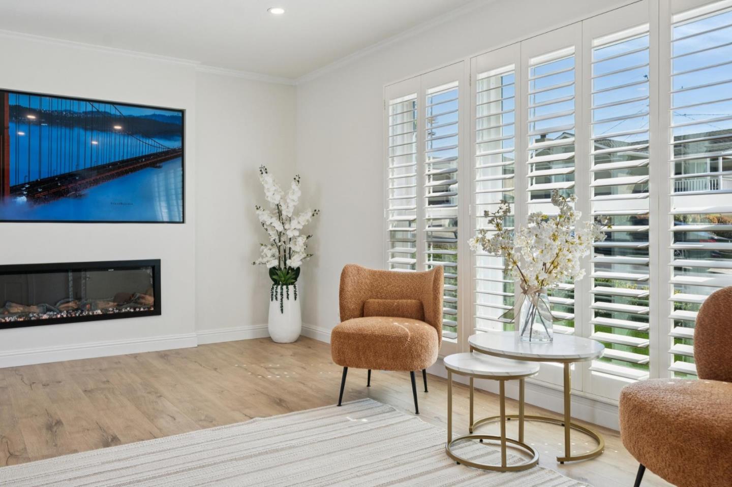 Bright living room with plantation shutters, rust-colored chairs, and a wall-mounted fireplace.