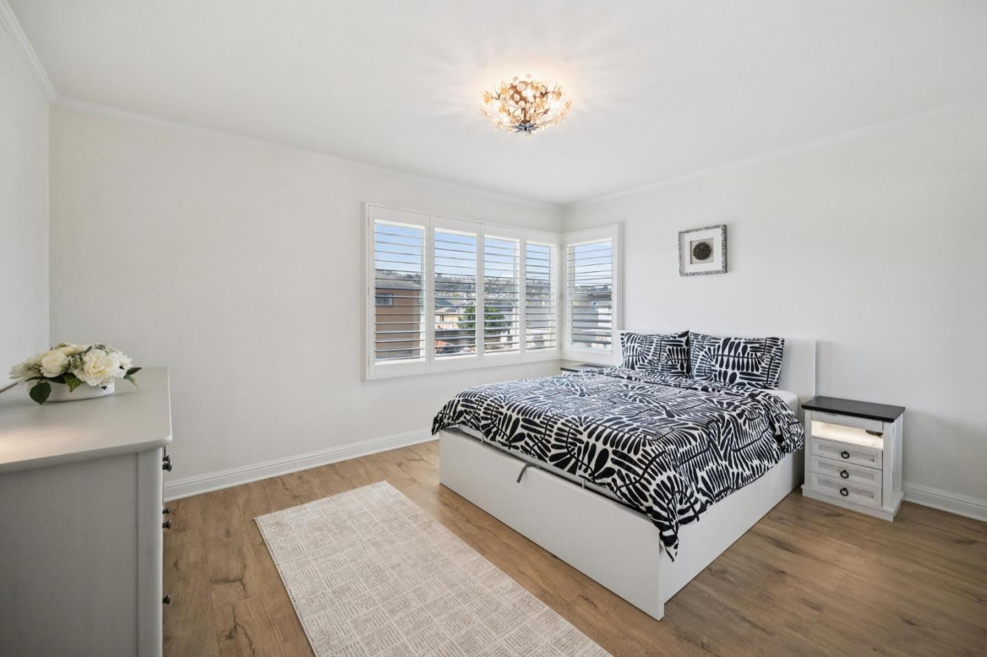 Staged bedroom with patterned bedding, bay windows with plantation shutters, and light wood flooring.