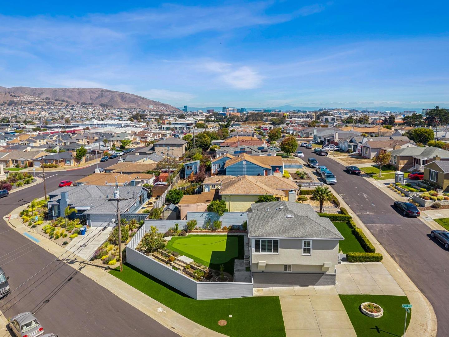 Aerial view of a corner home with a bright green yard, neighborhood streets, hills, and distant city buildings.